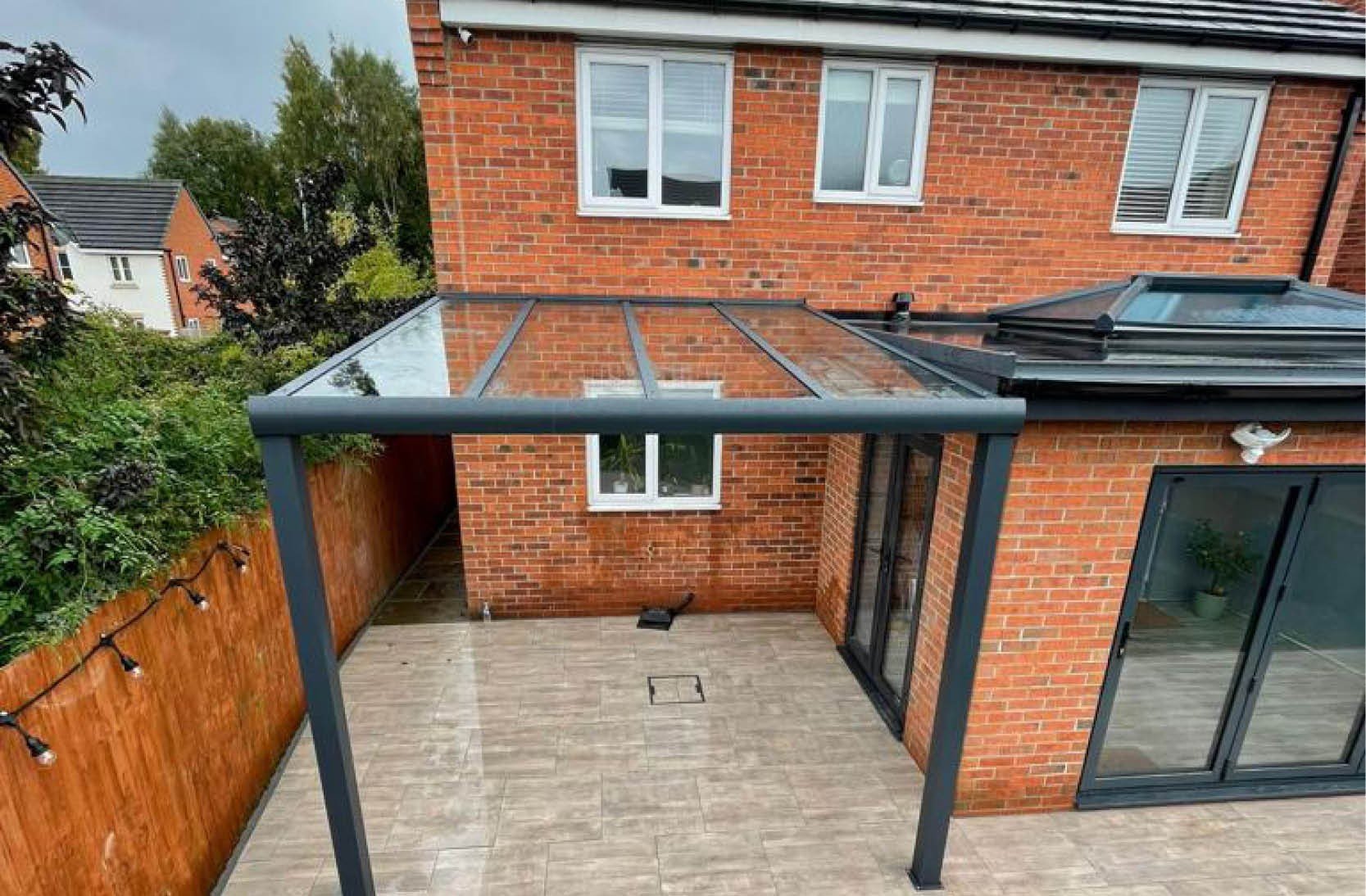A backyard patio area with a glass roof extension attached to a red brick house, surrounded by a wooden fence and trees.