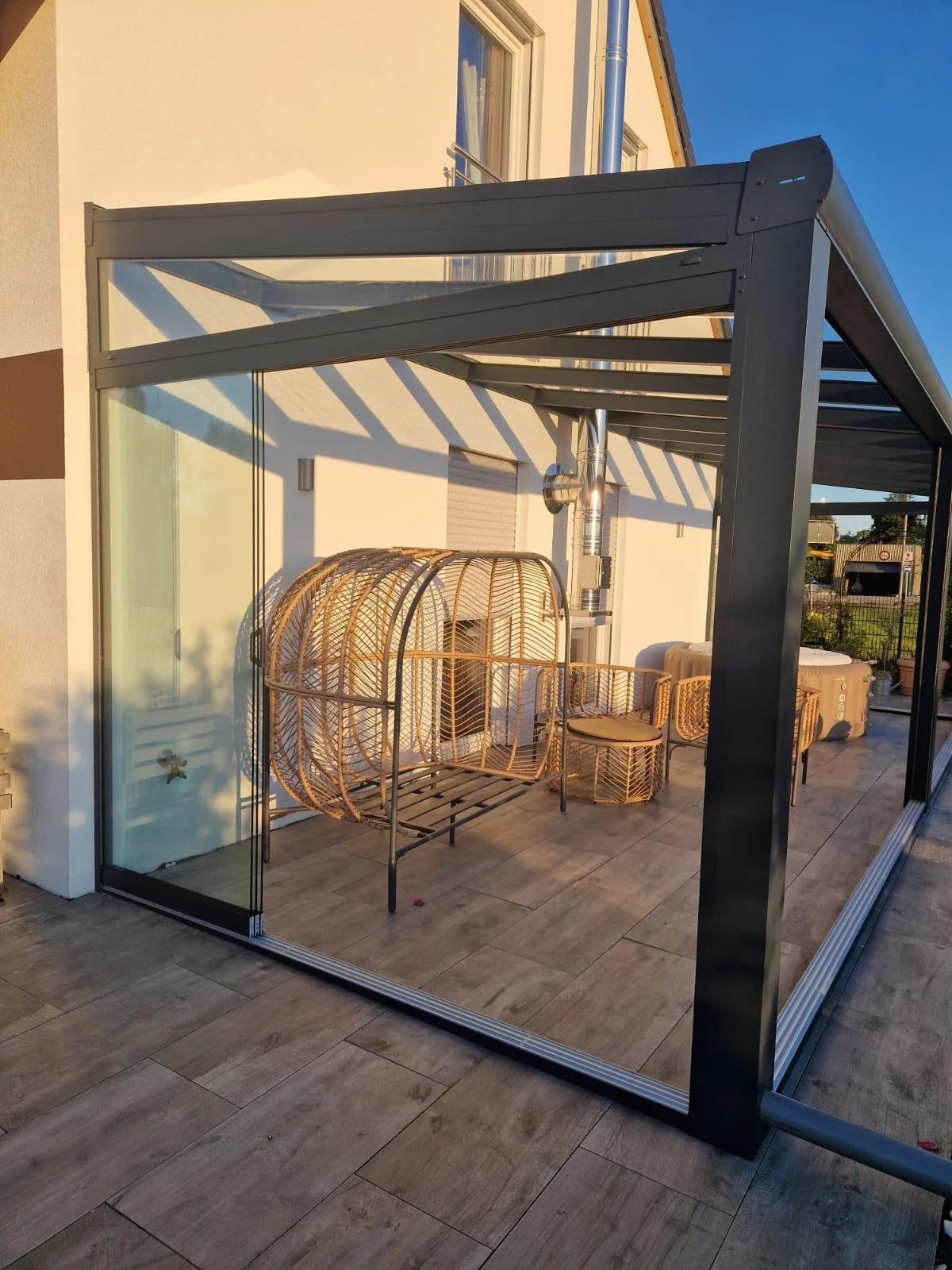 Outdoor patio area enclosed with a black metal and glass frame, with wicker furniture including a large hanging chair inside, set against a white wall with windows, and a blue sky above.