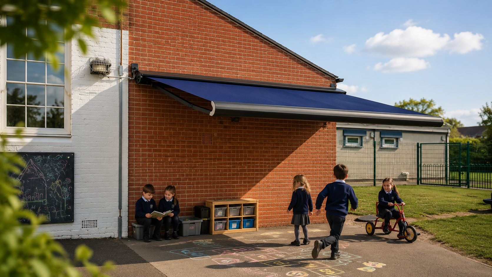 Children playing outside a school building with a hopscotch game on the pavement, two kids sitting and reading under the wall, while others run and ride a tricycle on the grass.