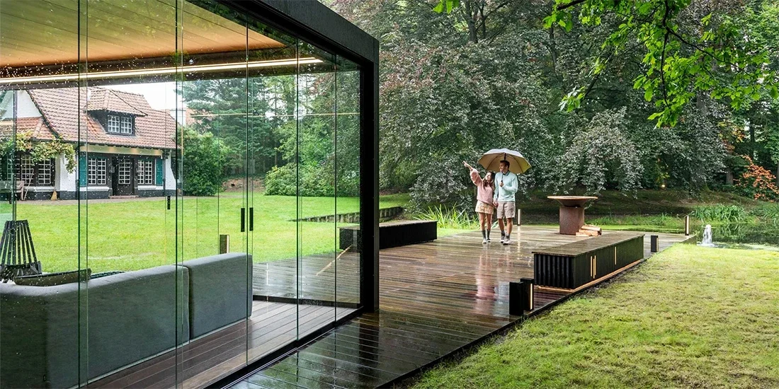 A couple walking on a wooden deck with an umbrella in a lush green backyard, with a modern glass house on the left and a large, flowering tree in the background.