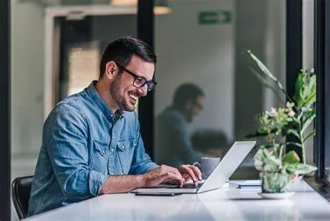 Homme souriant travaillant sur un ordinateur portable dans un bureau moderne avec des plantes et une grande fenêtre