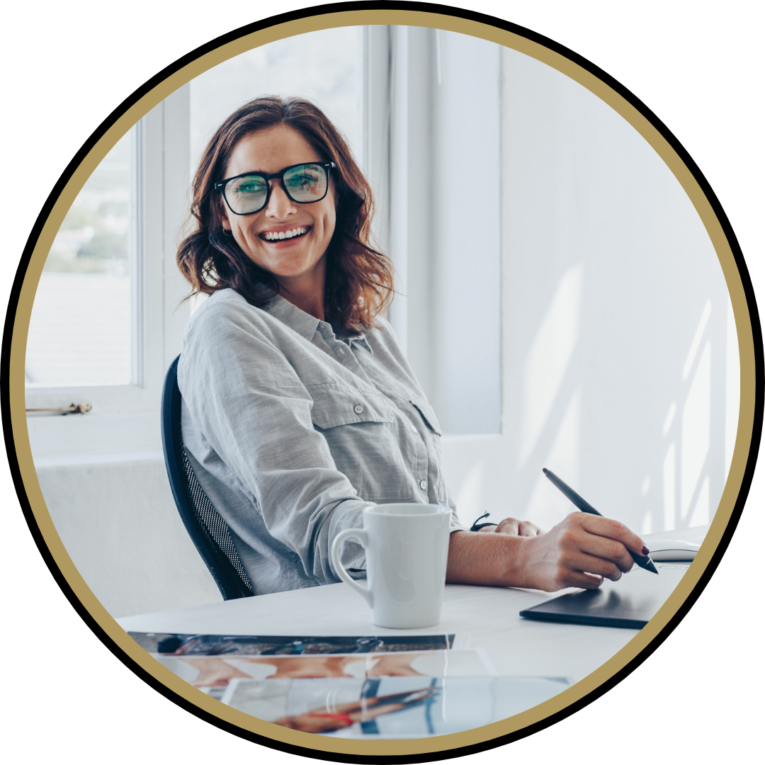 Femme souriante portant des lunettes assise à un bureau avec une tasse et une tablette, dans un bureau lumineux.