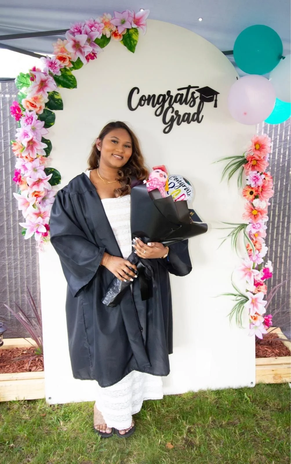 A woman in a graduation gown holding a bouquet of flowers and a diploma, standing in front of a decorated backdrop with a 'Congrats Grad' sign, pink and white flowers, and balloons.