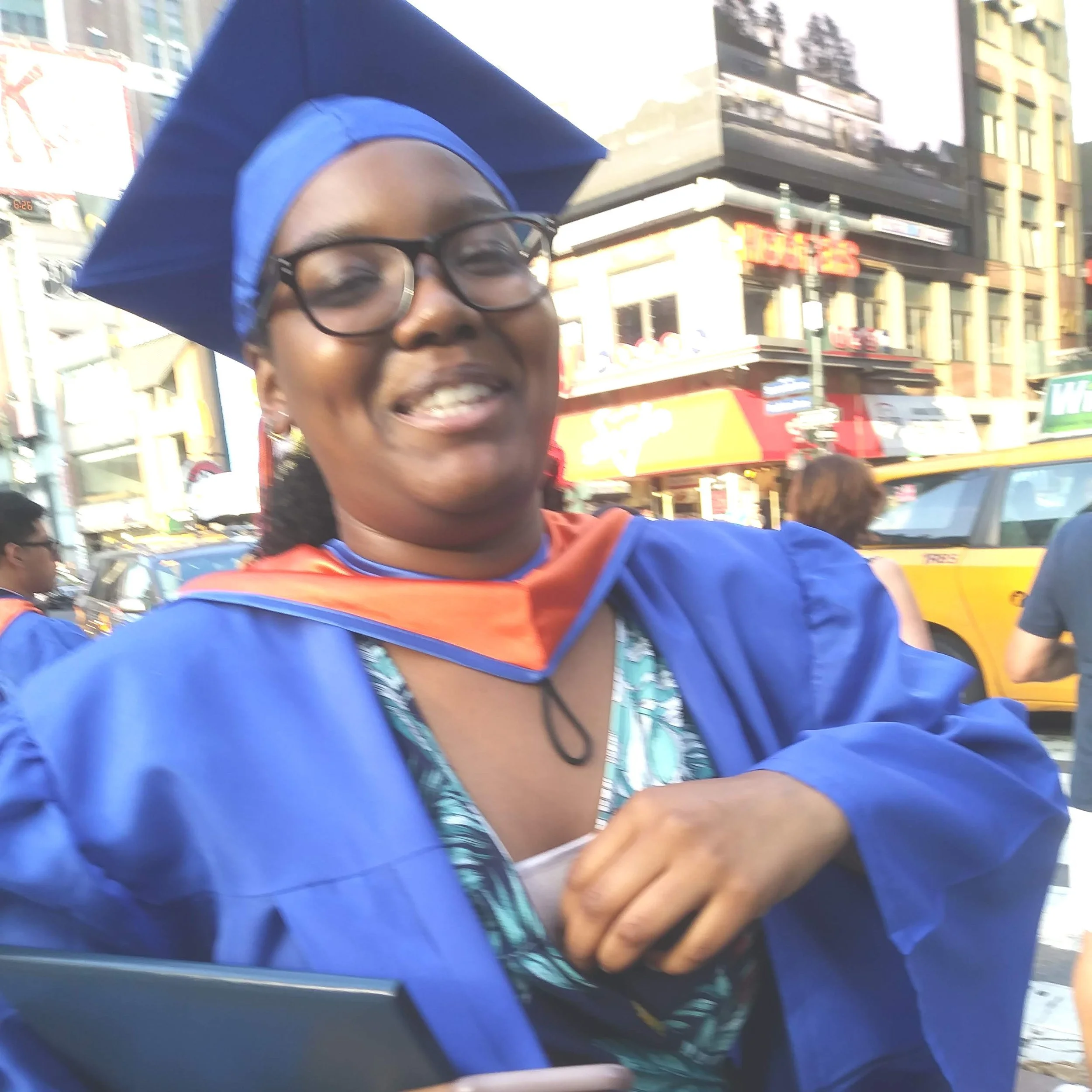A woman smiling at a graduation ceremony wearing a blue cap and gown with an orange collar, holding a phone in her hand, in an urban street with buildings, cars, and pedestrians in the background.