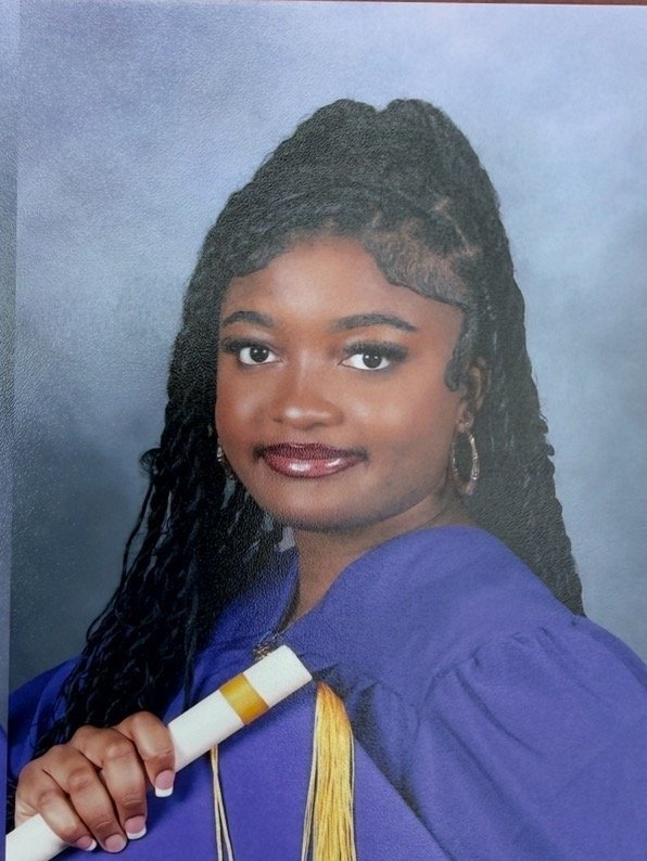 A young woman in a blue graduation gown holding a diploma, with long styled hair and hoop earrings, smiling in front of a neutral background.