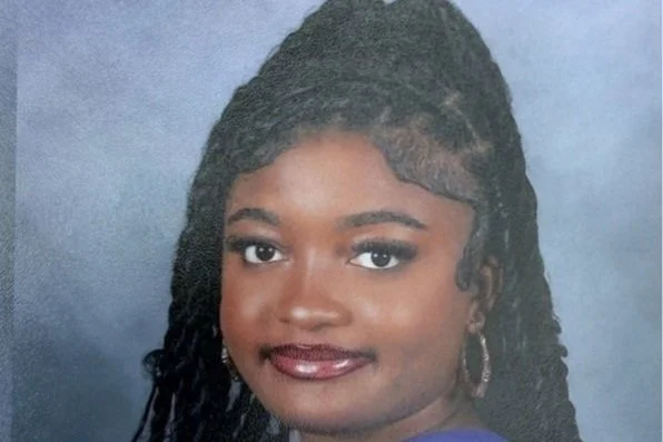 Portrait of a young woman with dark hair styled in twists, wearing hoop earrings, and smiling against a blue background.