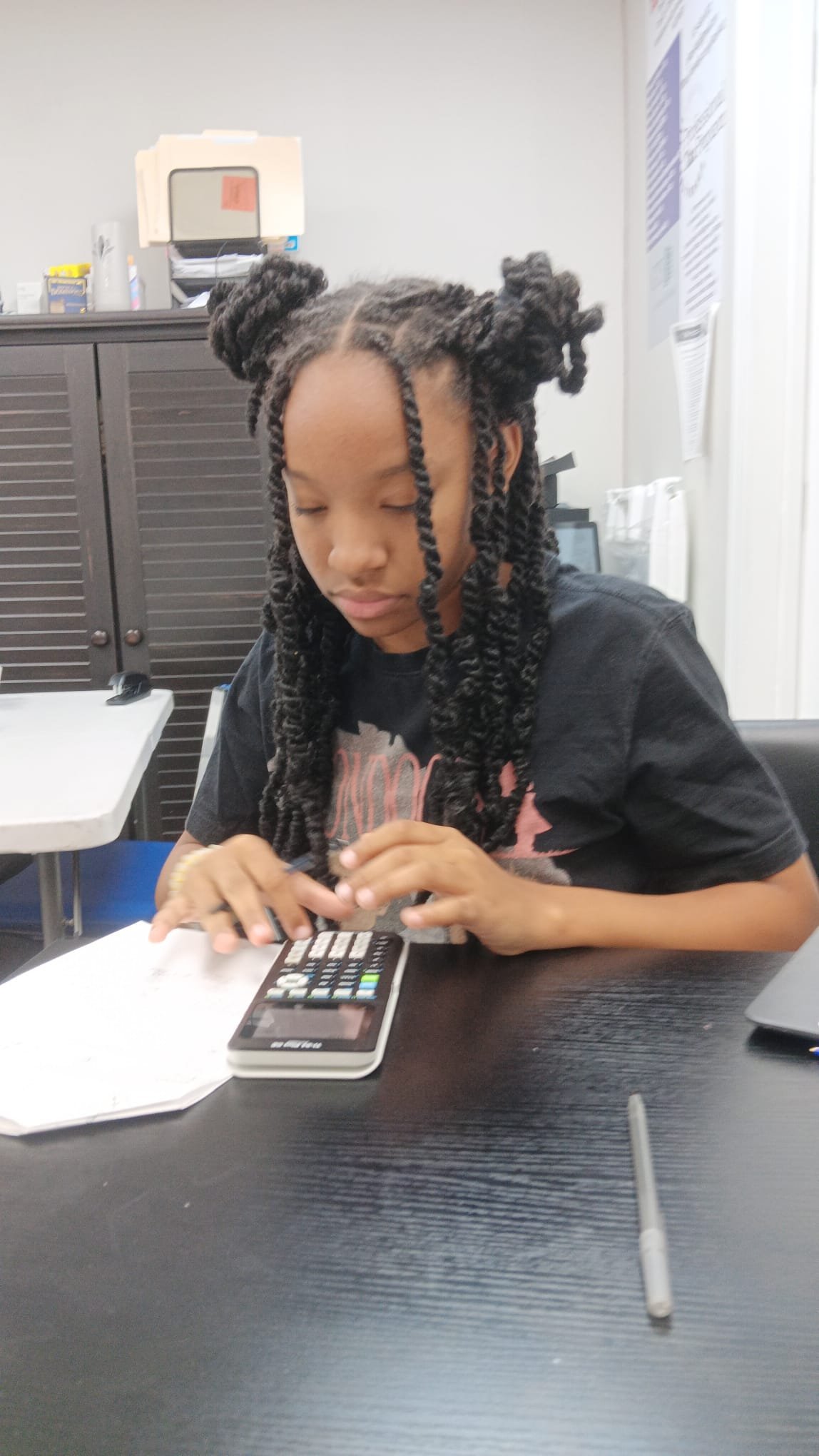 A young girl with styled twists in her hair is sitting at a table, focused on a calculator and a paper in an office or classroom setting.