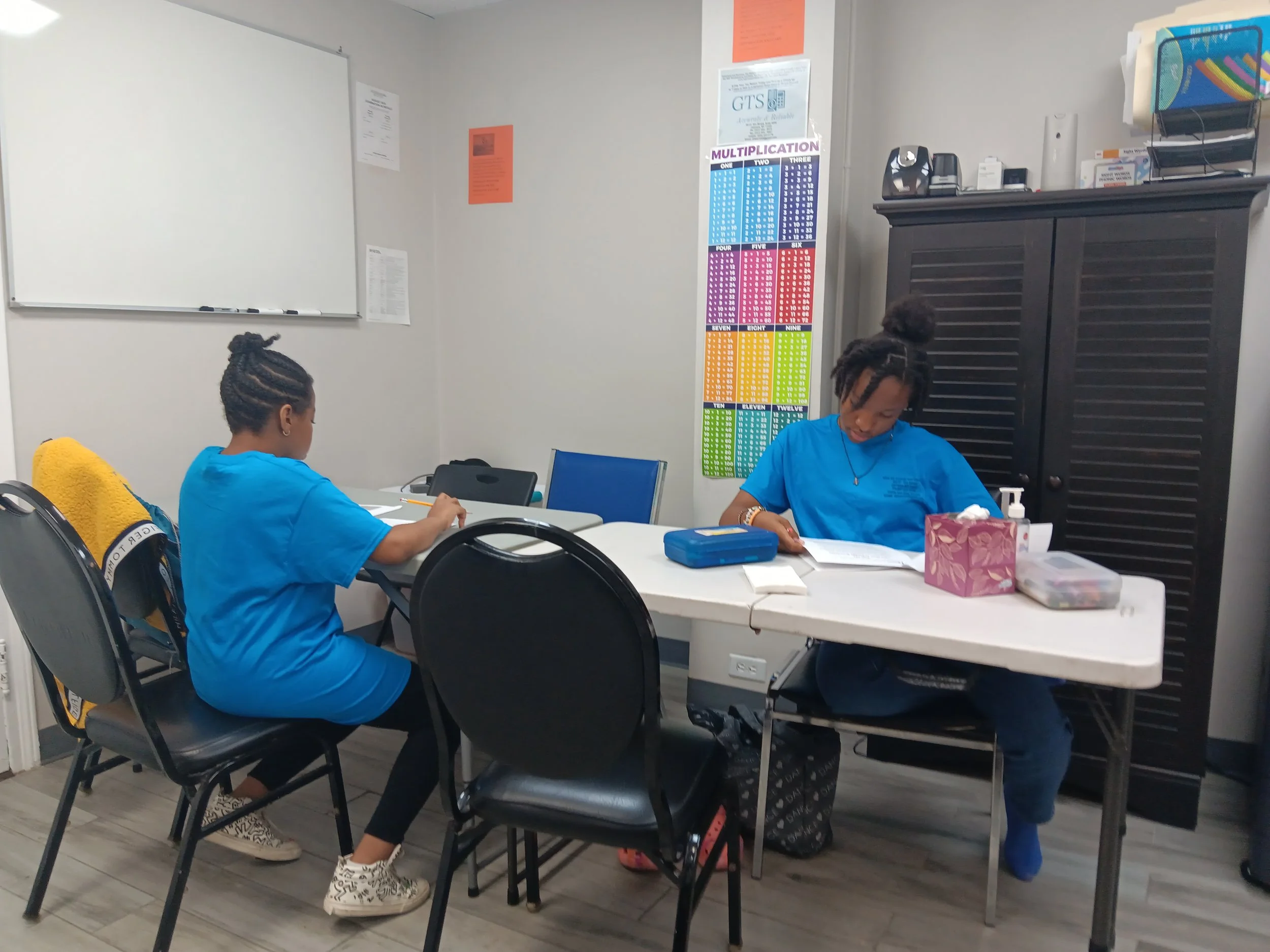 Two students in blue summer tshirts sitting at a table in the classroom, one writing and the other reading a book, with supplies and a colorful multiplication chart on the wall behind them.