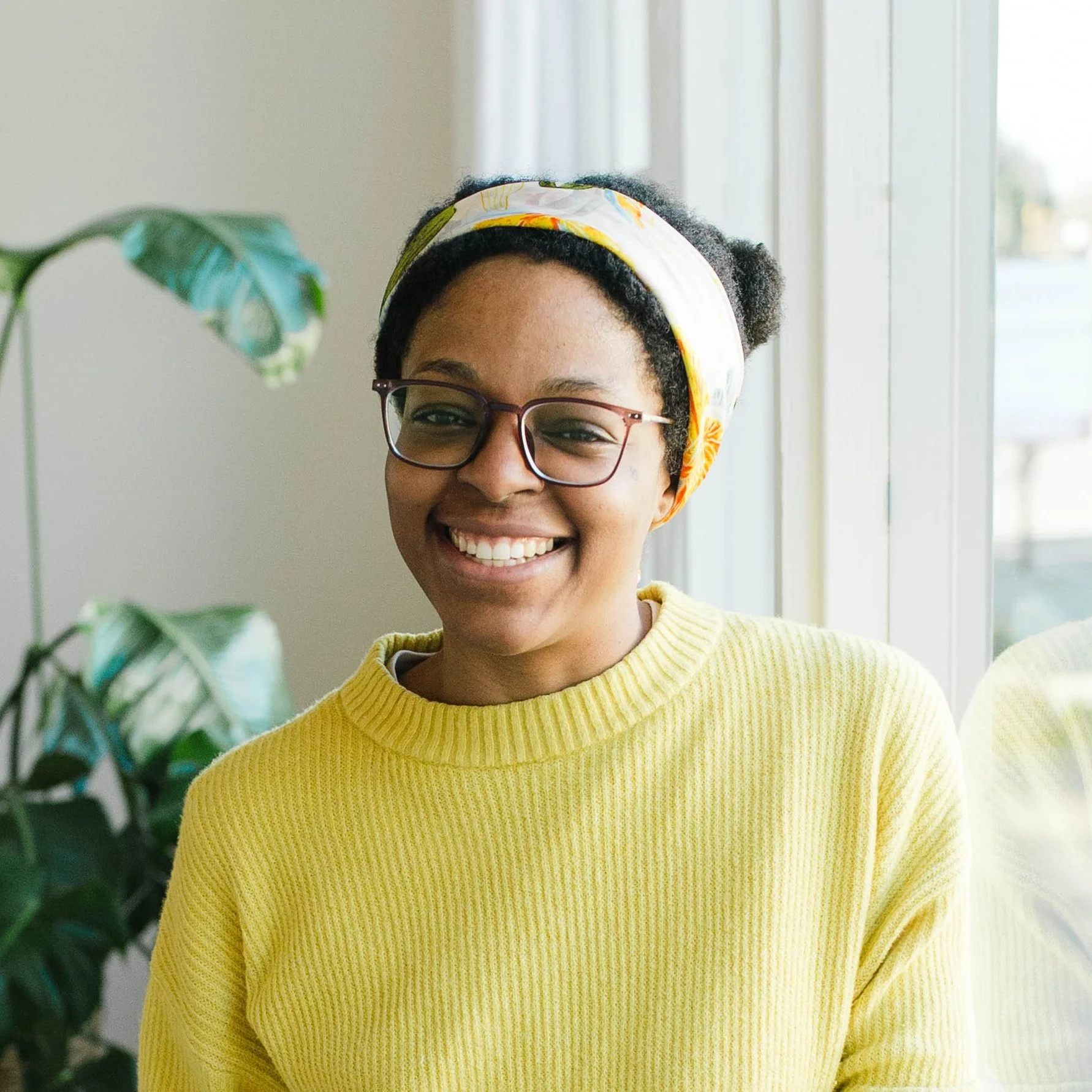 A woman sits in front of a window with a plant behind her. She's wearing a yellow scarf, brown-framed glasses, and a yellow sweater.