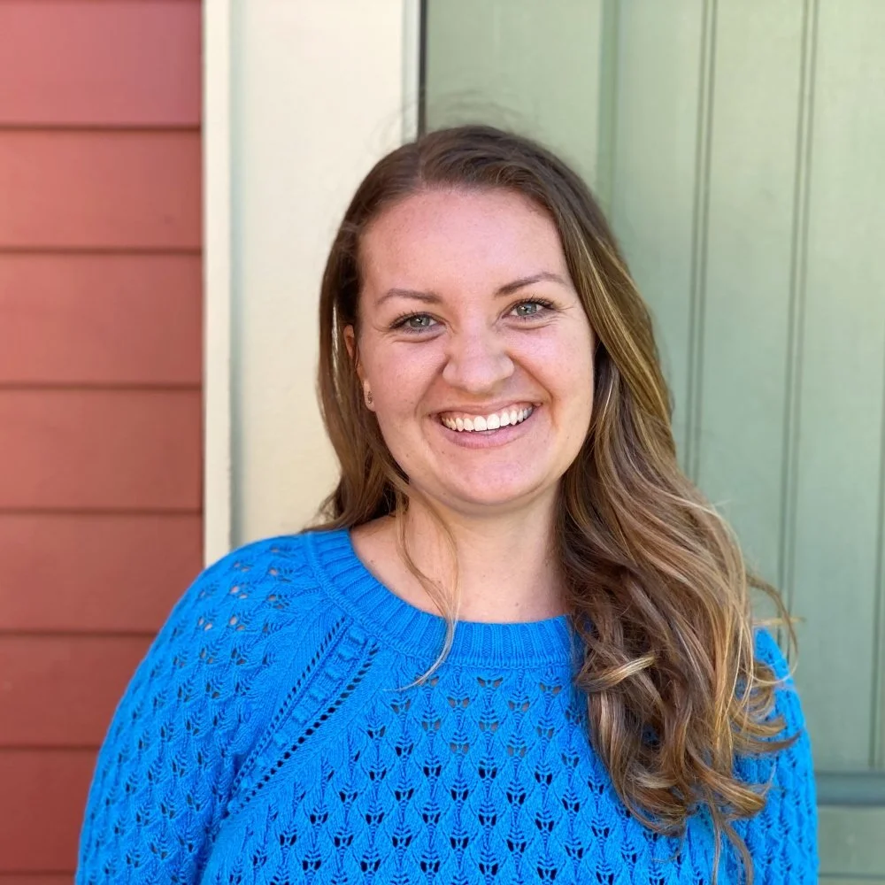 A woman with wavy brown hair wears a bright blue sweater. She stands in front of a brick-red and sage-green building.
