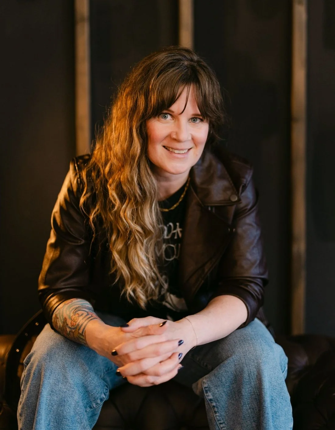 A woman leans forward with her arms clasped. She has long, wavy brown hair and is wearing a leather jacket, black tee, and light jeans. She sits in front of a dark backdrop.