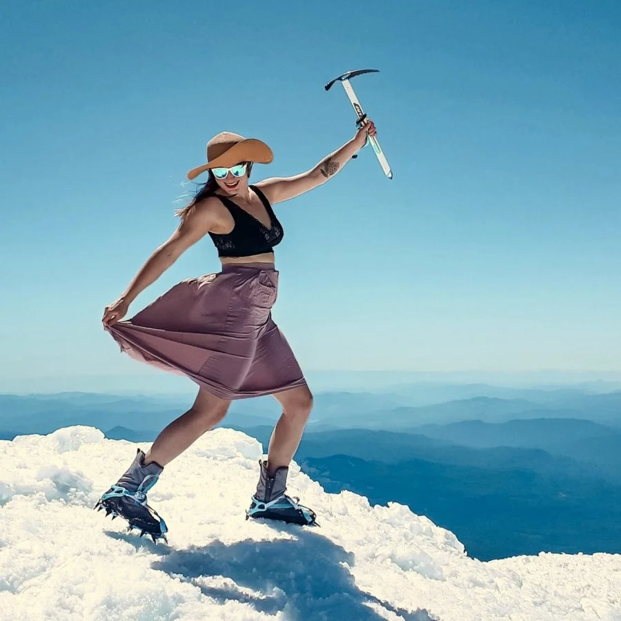 A woman on a snowy mountain summit holds an ice pick over her head. She's wearing a black sports bra, purple skirt, and mountaineering boots with crampons.