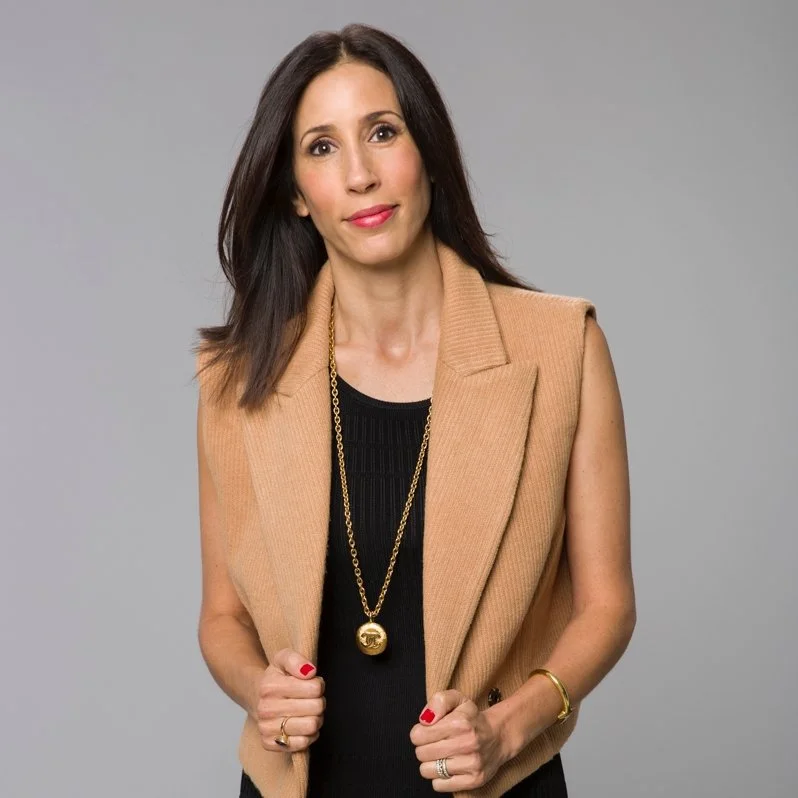 A woman wearing a black dress and tan vest, with dark brown hair, stands in front of a grey backdrop.