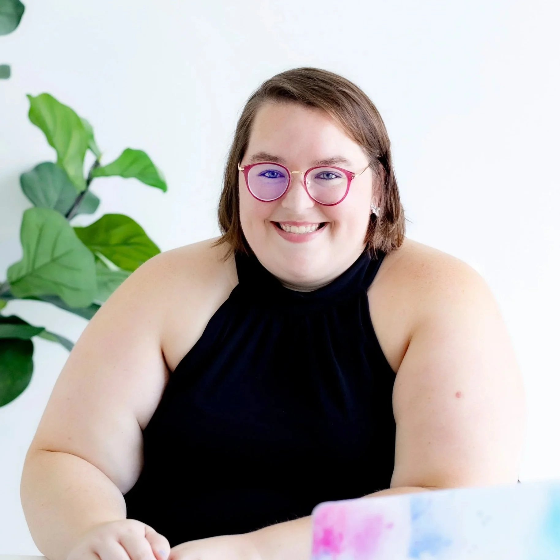 A woman with brown, bobbed hair and red-framed glasses wears a black sleeveless top. There is a green plant behind her, along with a white wall.