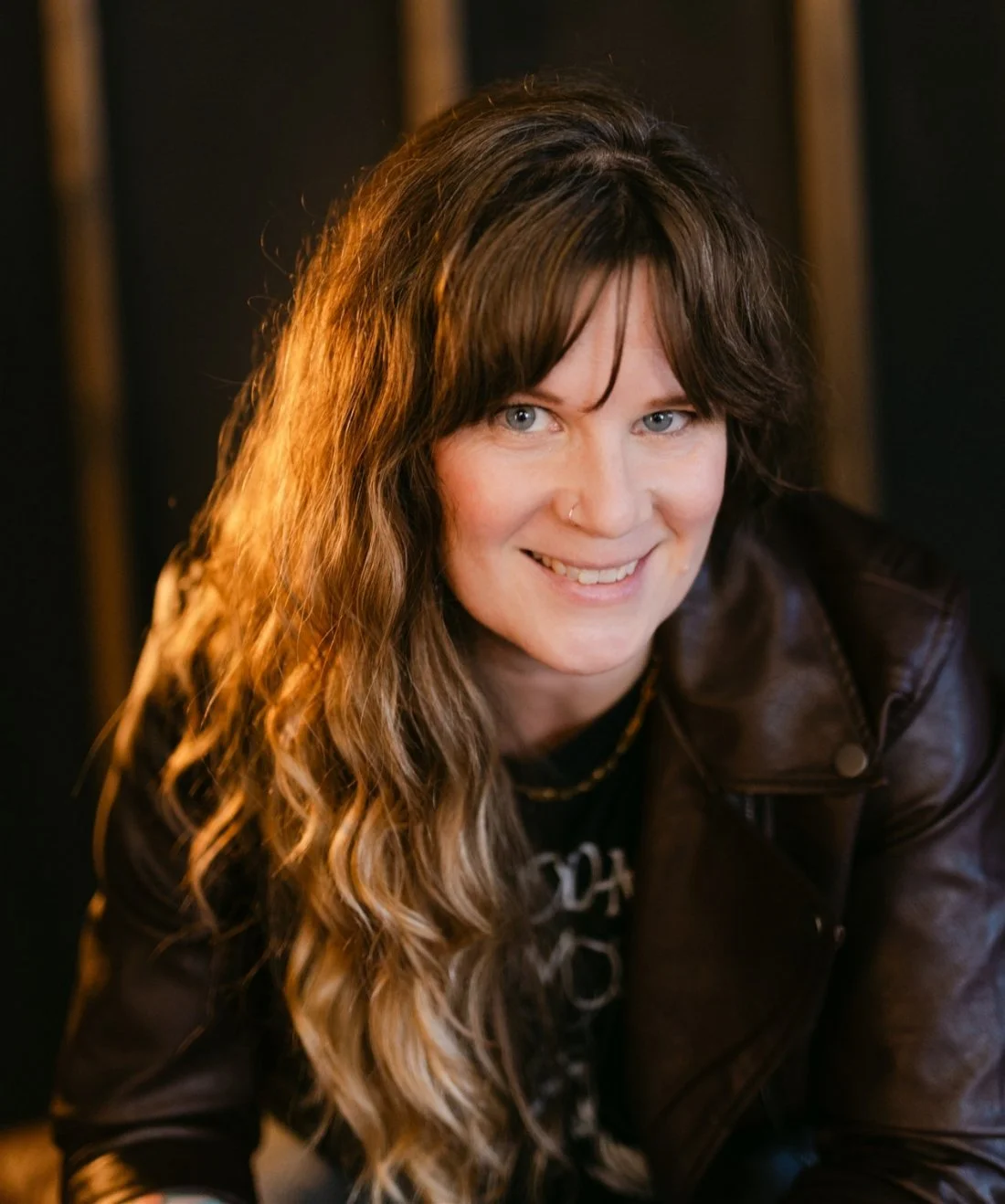 A woman wearing a leather jacket and black tee, with long brown wavy hair.