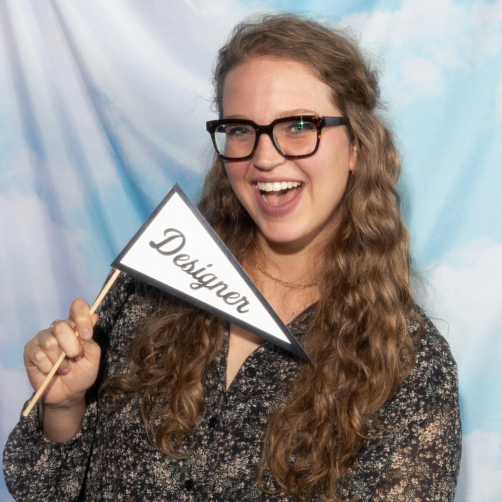A woman with long, wavy brown hair and dark-framed glasses sits in front of a cloud-printed fabric backdrop. She holds a small flag that says "Designer."