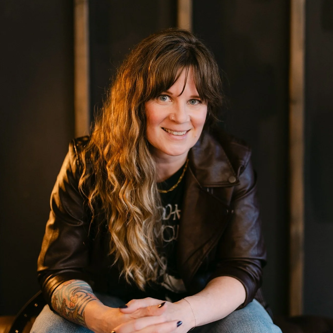 A woman with long, wavy brown hair wears a leather jacket and leans forward with her arms resting on her knees.