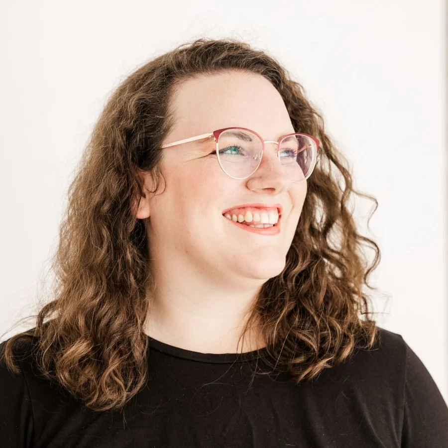 A woman with curly brown hair and gold-framed glasses wears a black shirt and smiles off camera.