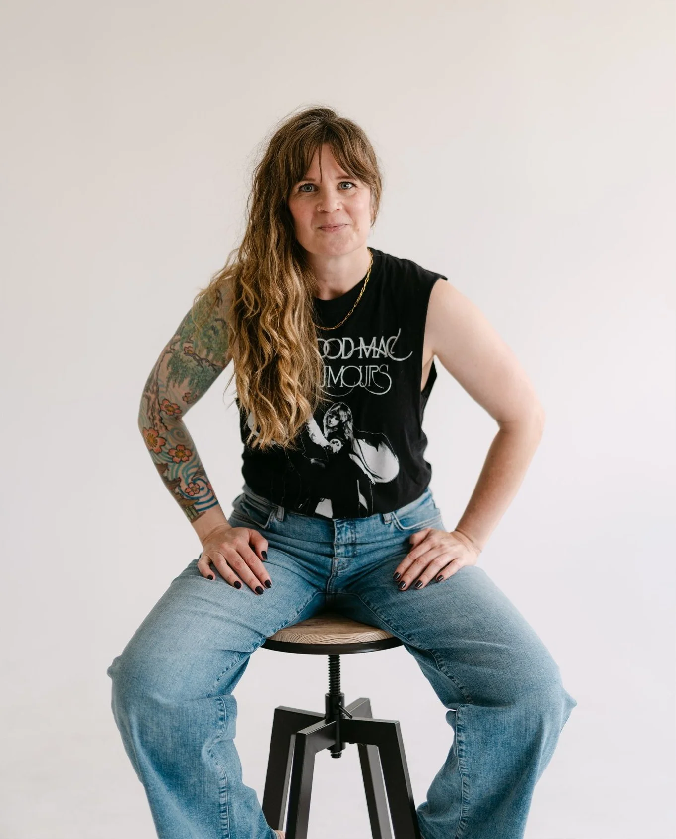 A woman with long, brown, wavy hair sits on a stool. She's wearing a black sleeveless top and light jeans, and has a tattoo covering one arm.