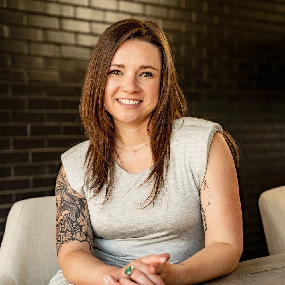 A woman with long, straight brown hair and an upper arm tattoo wears a grey top. She's sitting at a table with her arms resting on the tabletop.