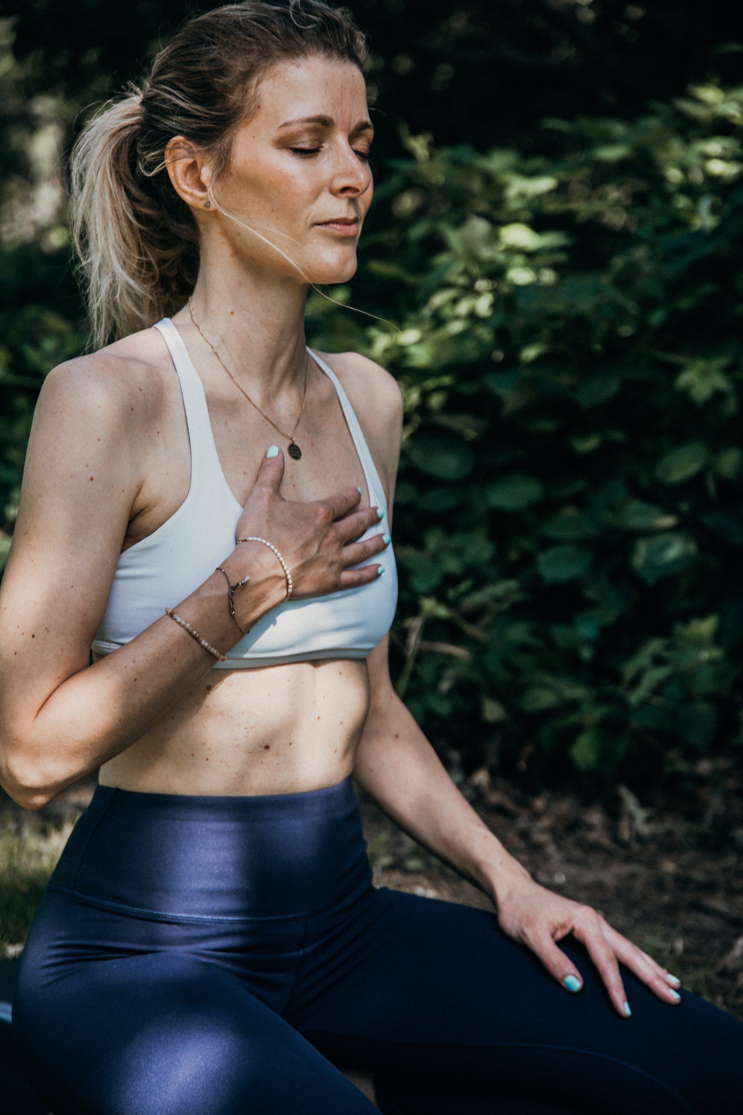Eine Frau macht Yoga im Grünen, sitzt in einem meditierenden Zustand mit geschlossenen Augen, trägt Sportbekleidung und Schmuck, umgeben von Büschen und Bäumen.