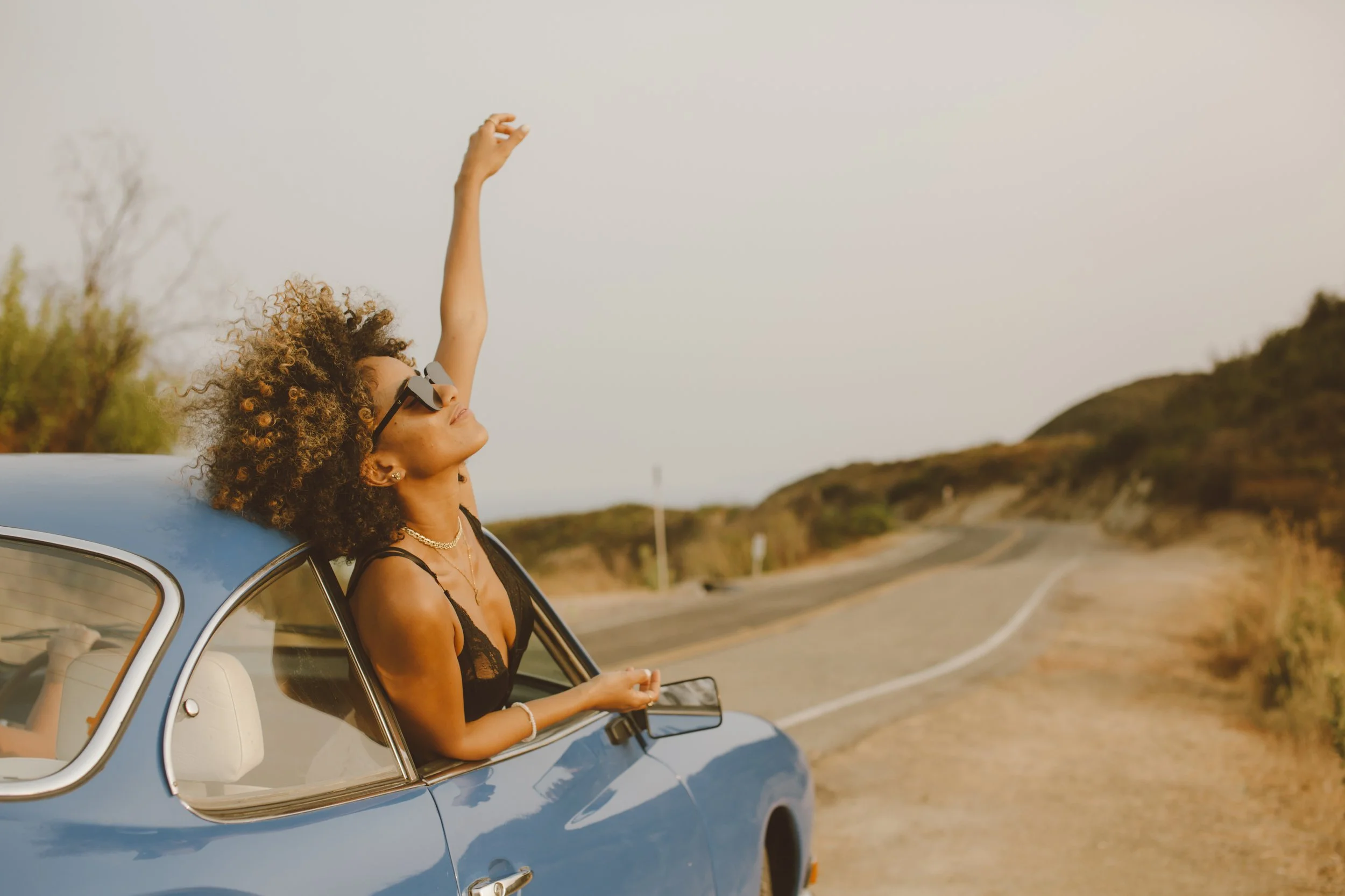 Woman with curly hair and sunglasses leaning out of a blue vintage car window, stretching her arm up, on a scenic desert road.