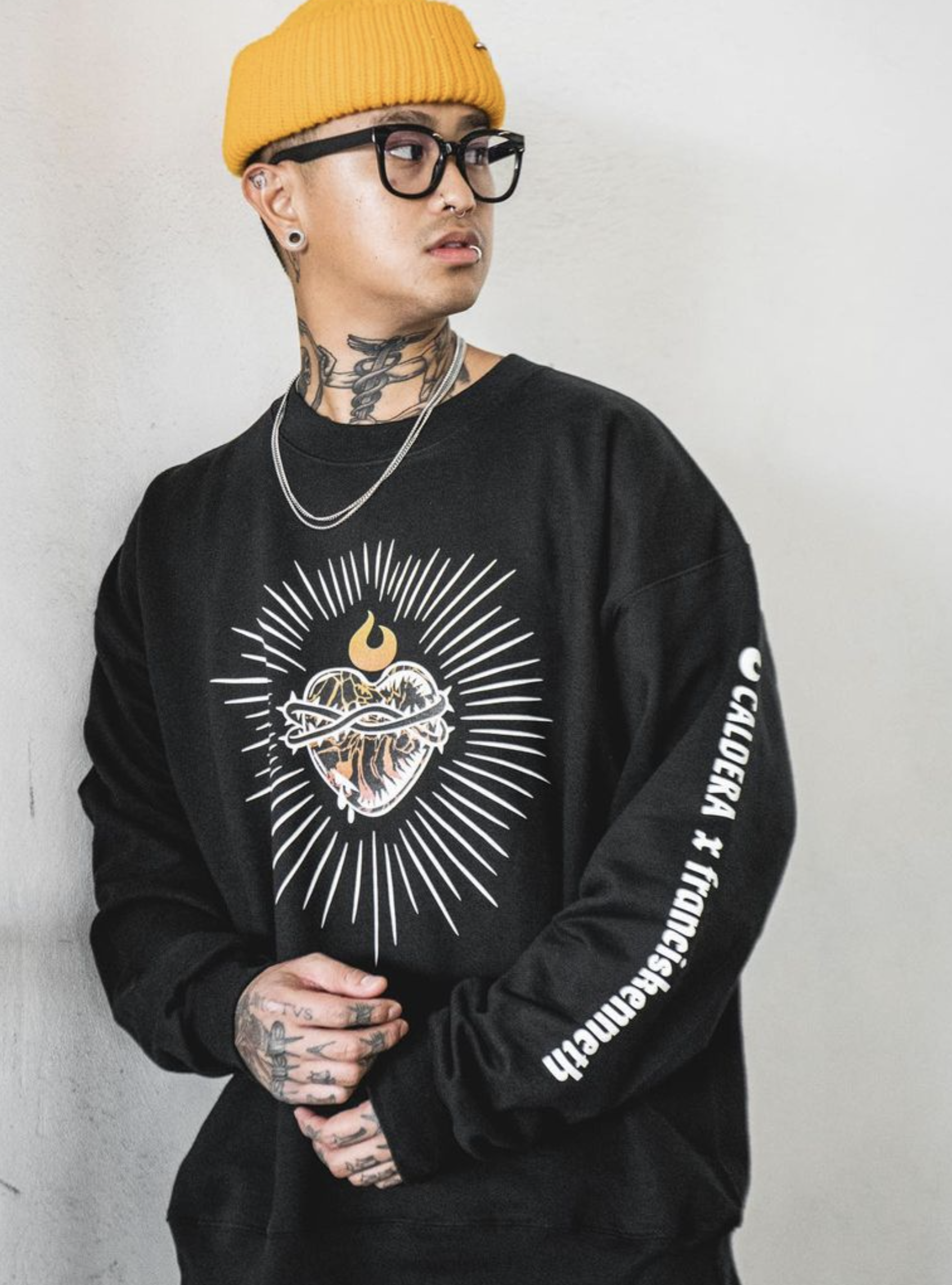 A young person with tattoos, earrings, glasses, and a nose piercing, wearing a yellow beanie and black sweatshirt with white text and graphic art, standing against a plain wall.