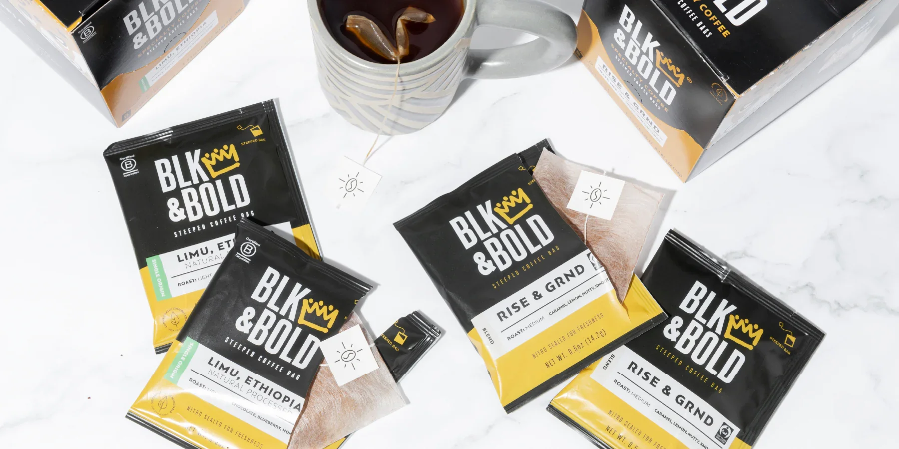 Assorted packs of BLK & BOLD Stepped Coffee Bags, a mug with coffee, and a box of coffee on a white marble surface.
