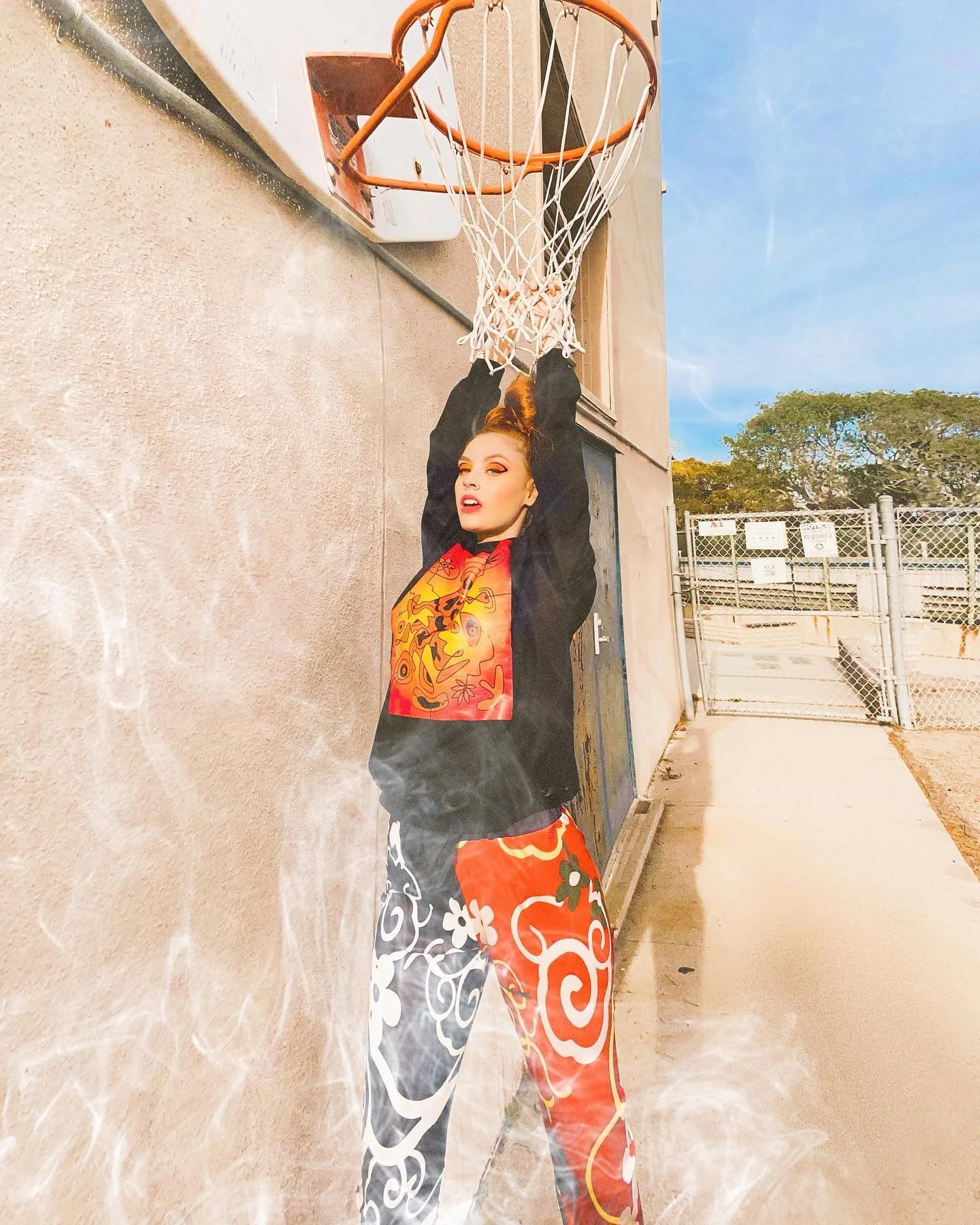 A woman hanging from a basketball hoop with her arms raised above her head, outdoors against a building wall, with a fence and trees in the background.