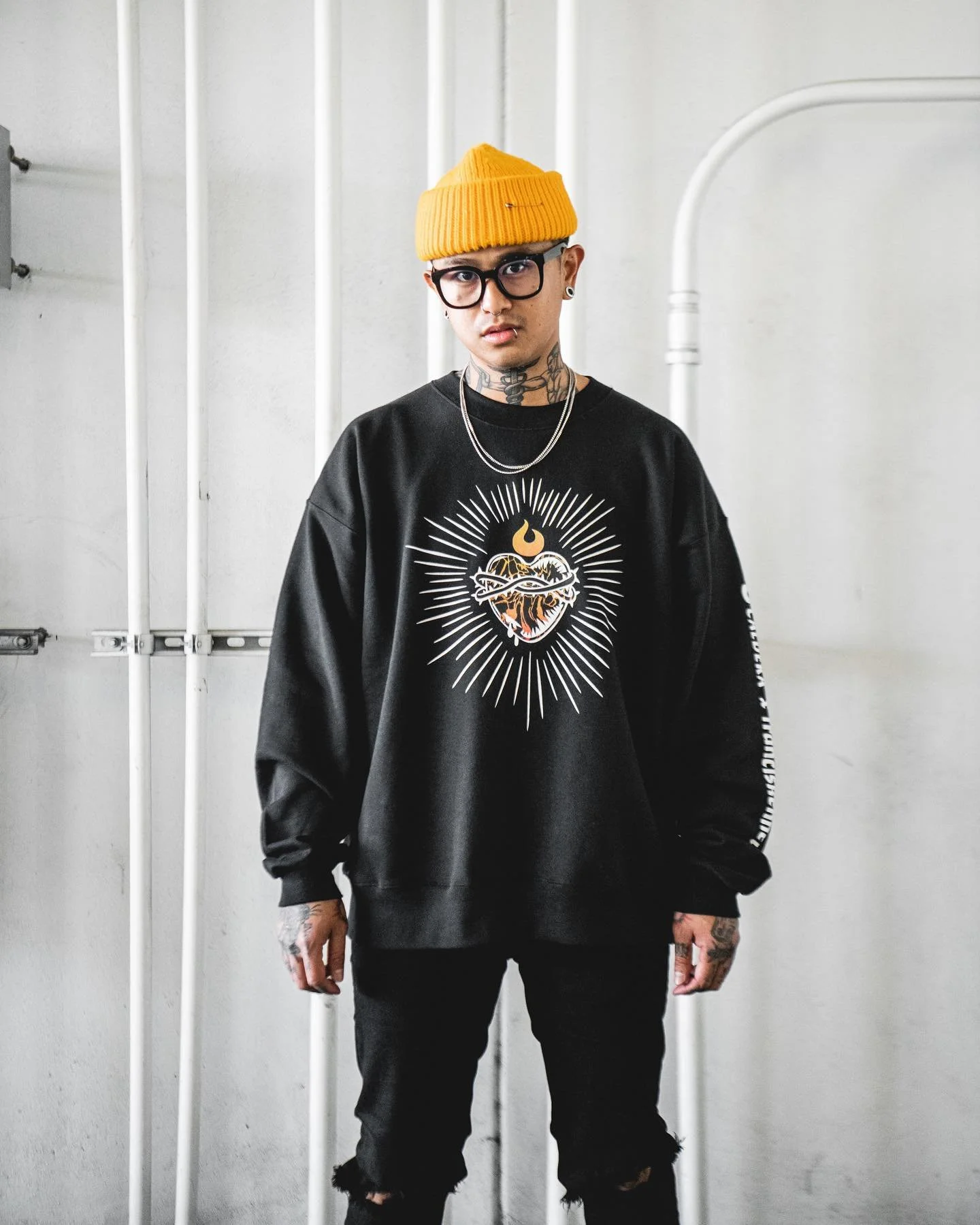 A young man with tattoos, wearing glasses, a yellow beanie, and black ripped jeans, standing against a white wall with pipes, dressed in a black sweatshirt with a graphic of a heart and lightning, and holding a silver chain.