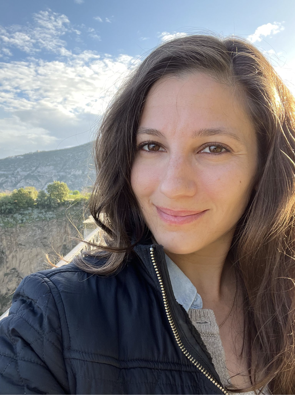 A woman with long brown hair taking a selfie outdoors during daytime, with mountains, trees, and a partly cloudy sky in the background.