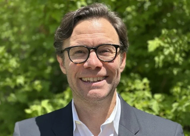 A man wearing glasses and a suit smiling outdoors with green trees in the background.