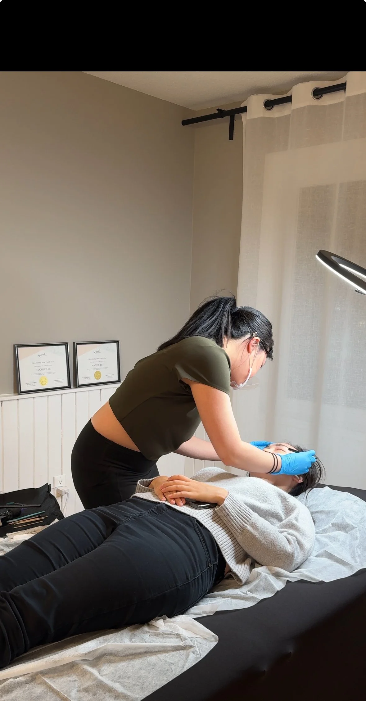 A woman wearing a face mask and gloves is performing a beauty treatment on a client lying on a table in a clinic room. There are framed certificates on the wall and a lamp providing focused light on the client's face.
