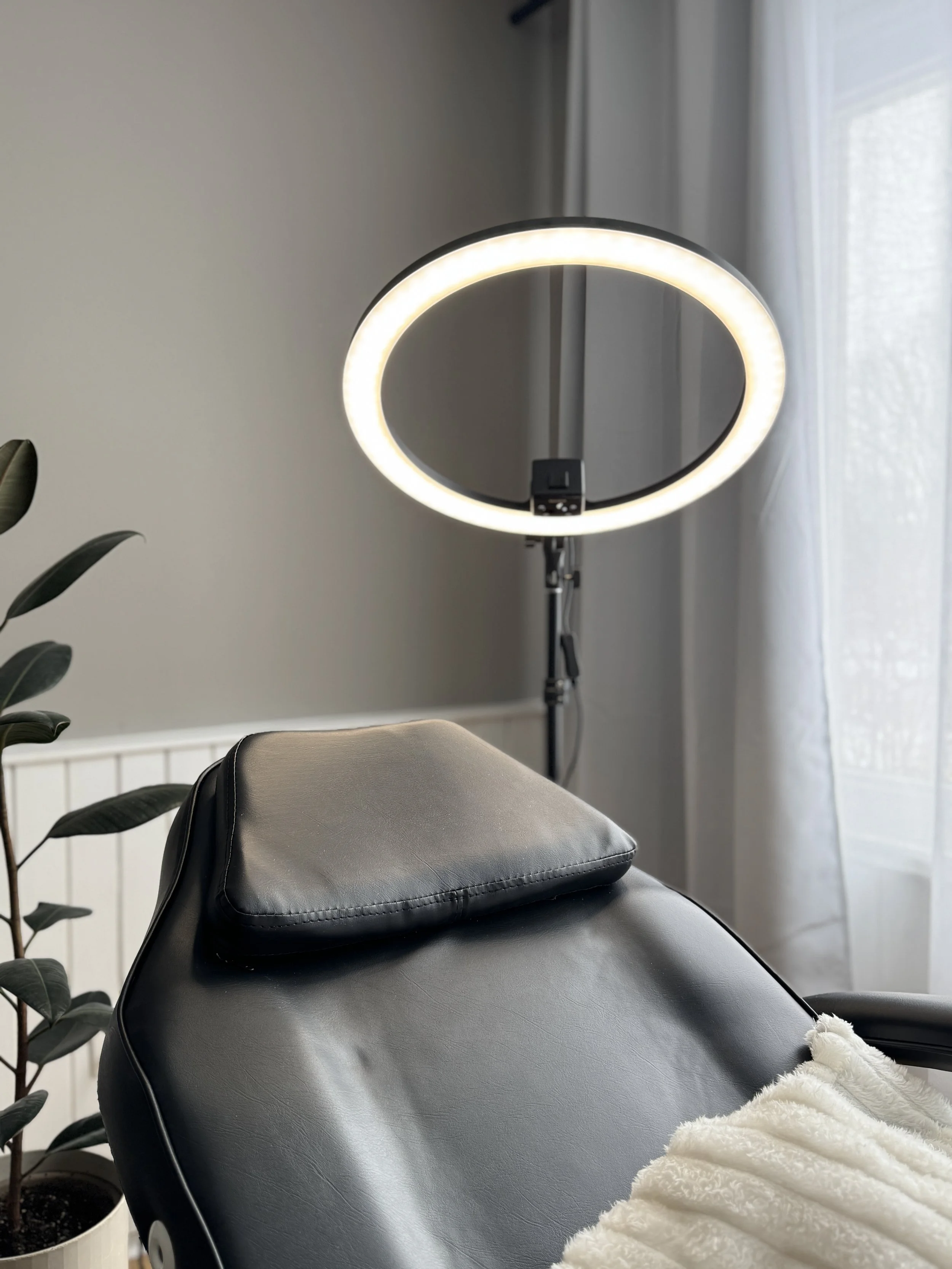 Professional beauty salon setup with black leather chair, ring light, and potted plant in a minimalistic room.