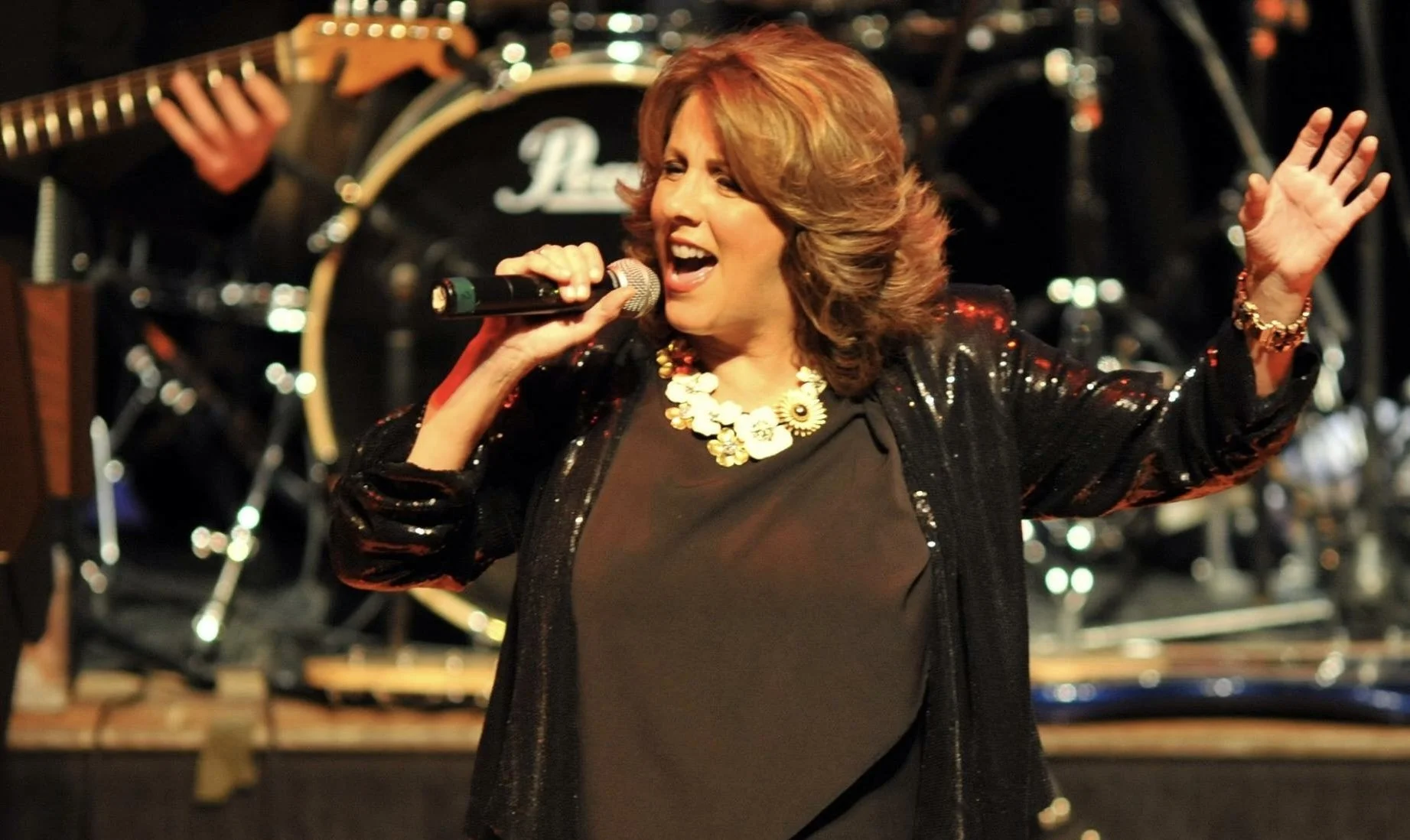 A woman singing into a microphone with a band in the background, wearing a black sequined jacket and a floral necklace.