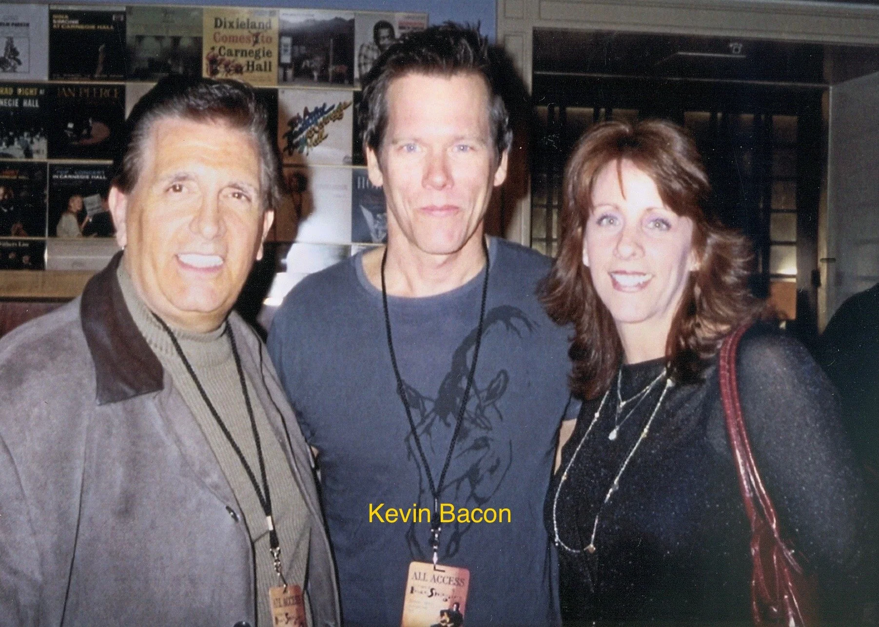 Three people, two men and one woman, standing close together indoors, smiling at the camera. The background features posters and signs, including one for a musical event. The man on the left has gray hair and wears a beige and brown jacket with a bad