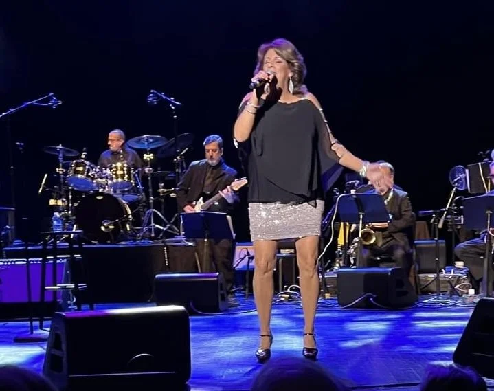 A woman singing on stage with a band in the background, holding a microphone, wearing a dark top and a shiny silver skirt, with stage equipment and musical instruments around her.