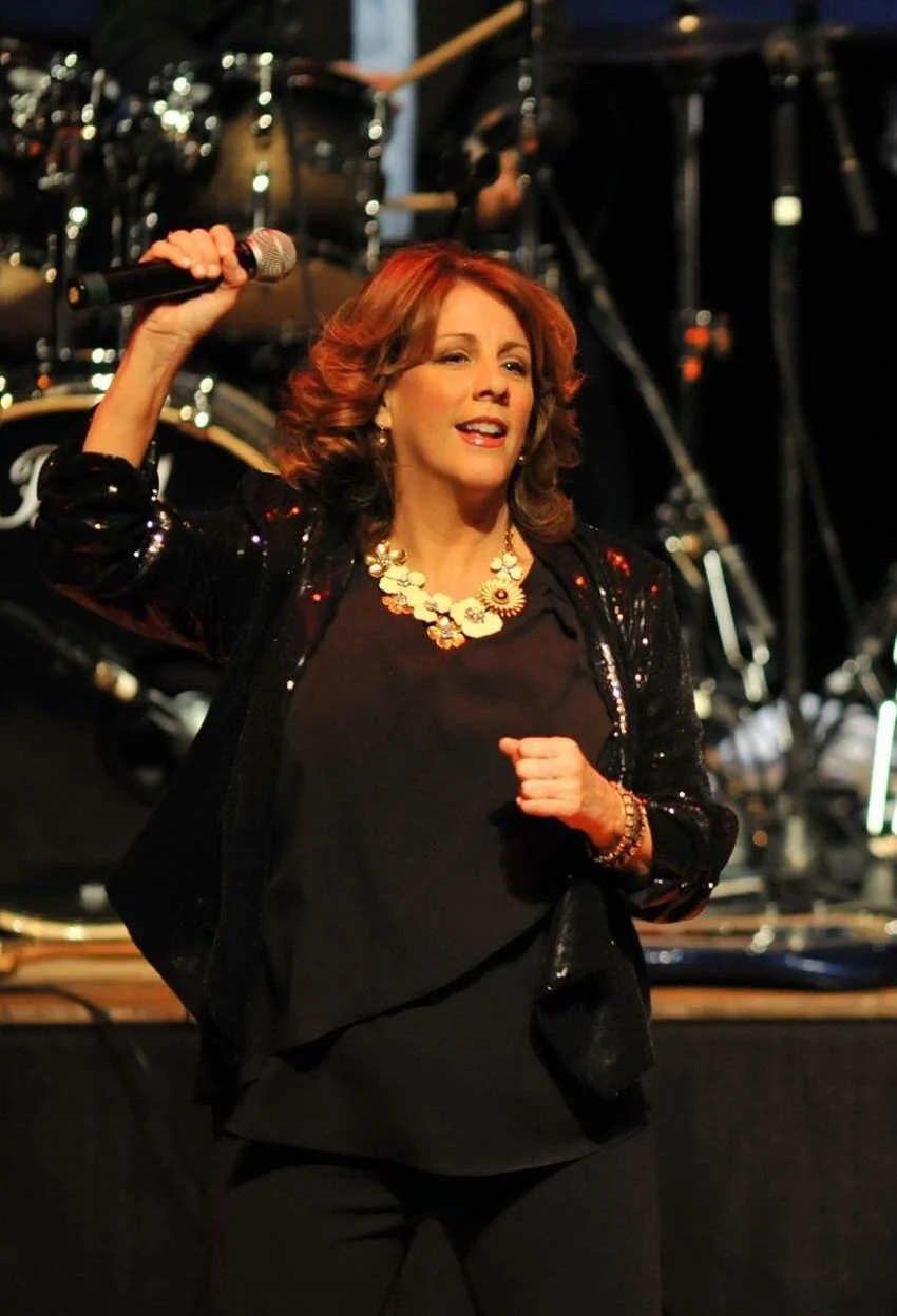 A woman with reddish-brown curly hair singing into a microphone on stage, wearing a black sequined jacket and a floral necklace, with musical instruments and stage equipment in the background.
