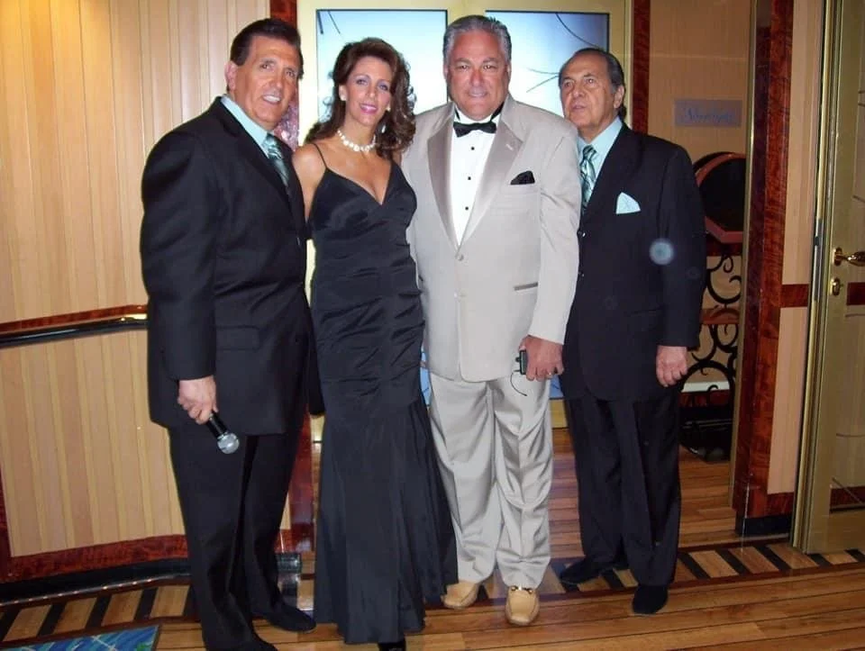 Four adults dressed in formal attire posing together indoors, with a wooden wall and screens in the background.