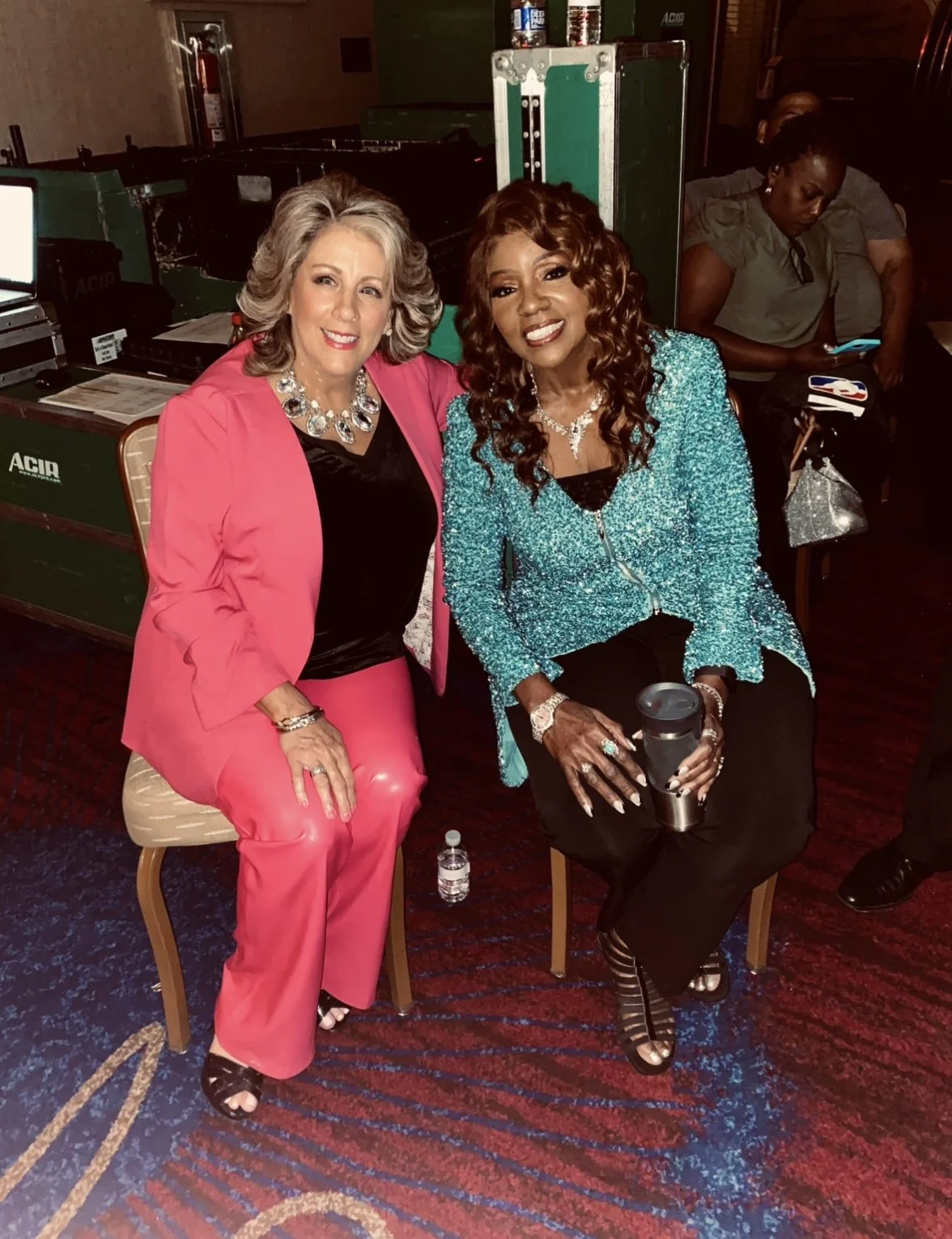 Two women sitting and smiling at a social event, with a speaker and a woman using her phone in the background, in a dimly lit room with patterned carpet.