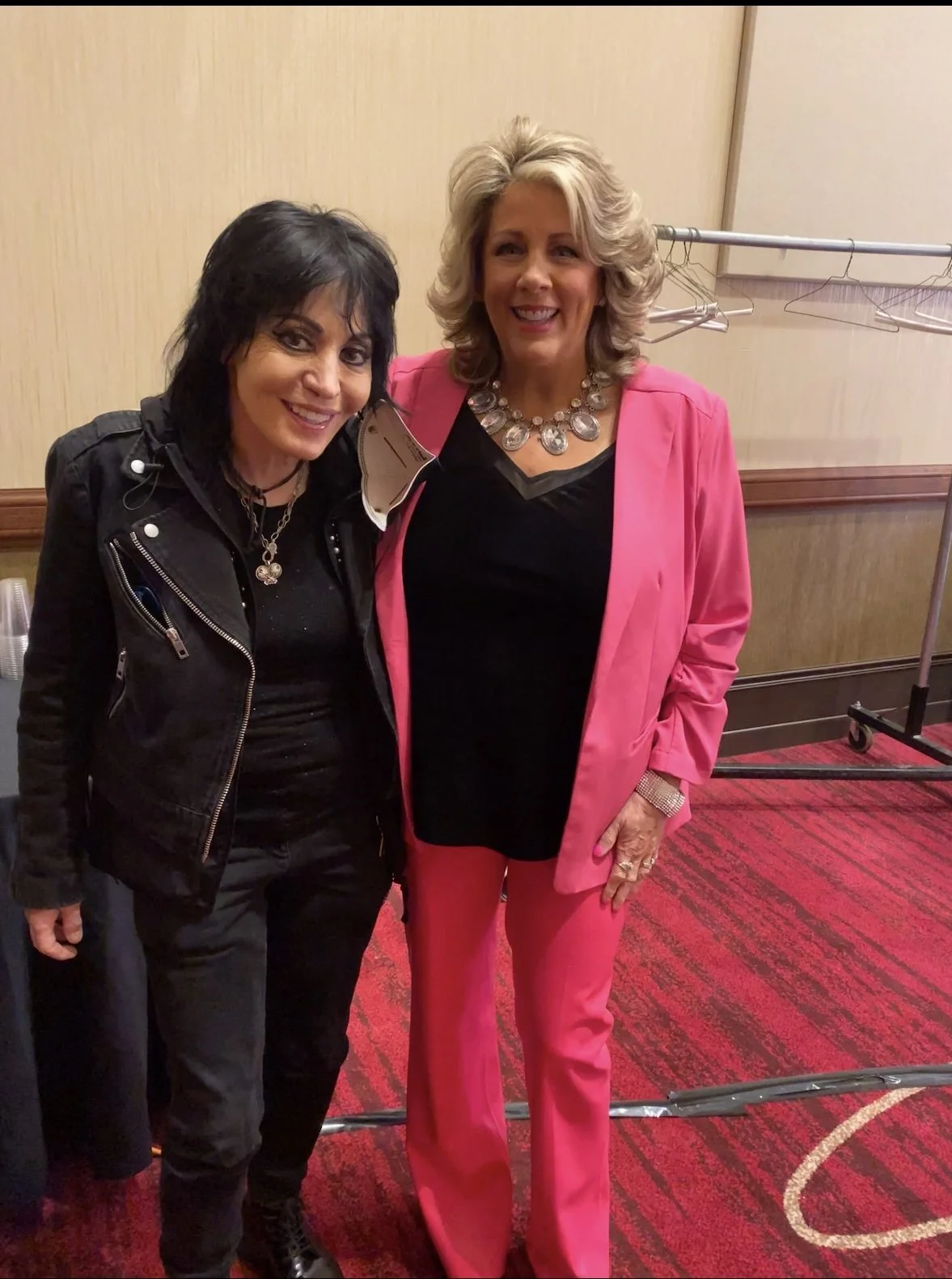 Two women standing together, smiling for the camera, indoors with a beige wall and red patterned carpet in the background.
