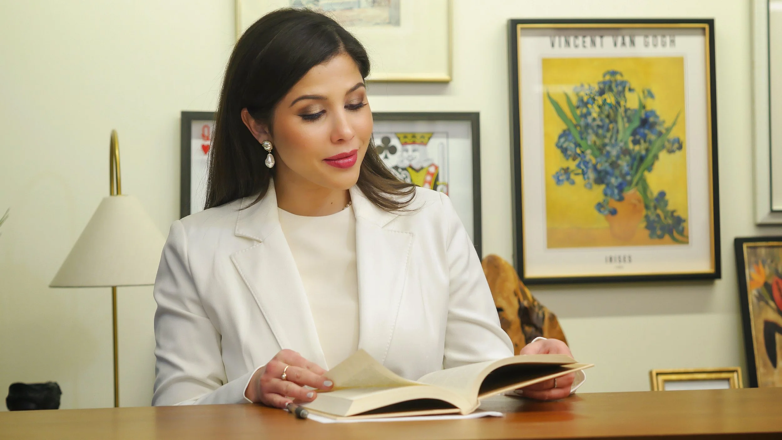 A woman with dark hair, wearing a white blazer and pearl earrings, sitting at a desk and reading a book in an art-filled room.