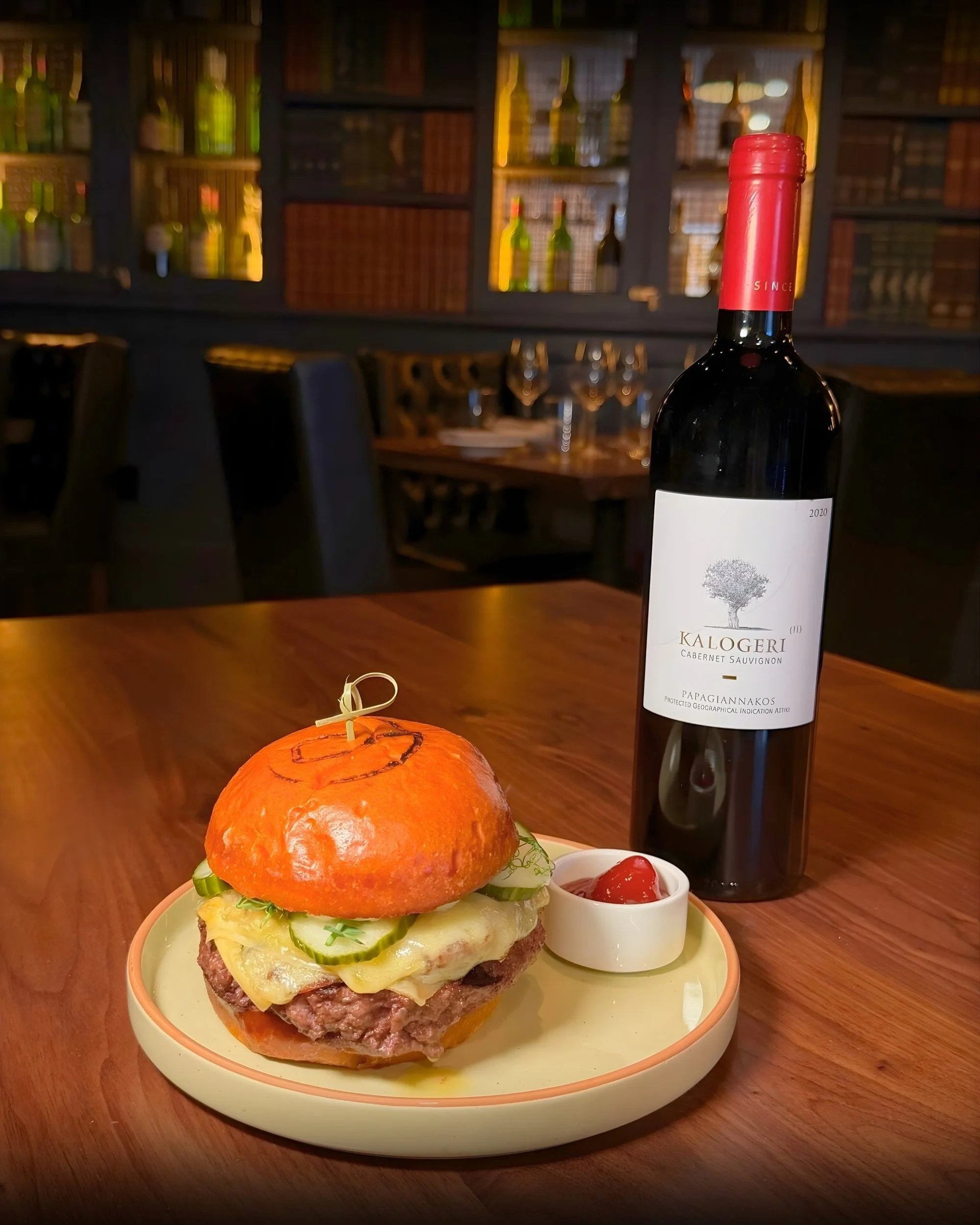 A burger with cucumber slices and melted cheese on a bun, served on a plate with a side of ketchup, a bottle of red wine, and a small dish of ketchup in a restaurant setting.