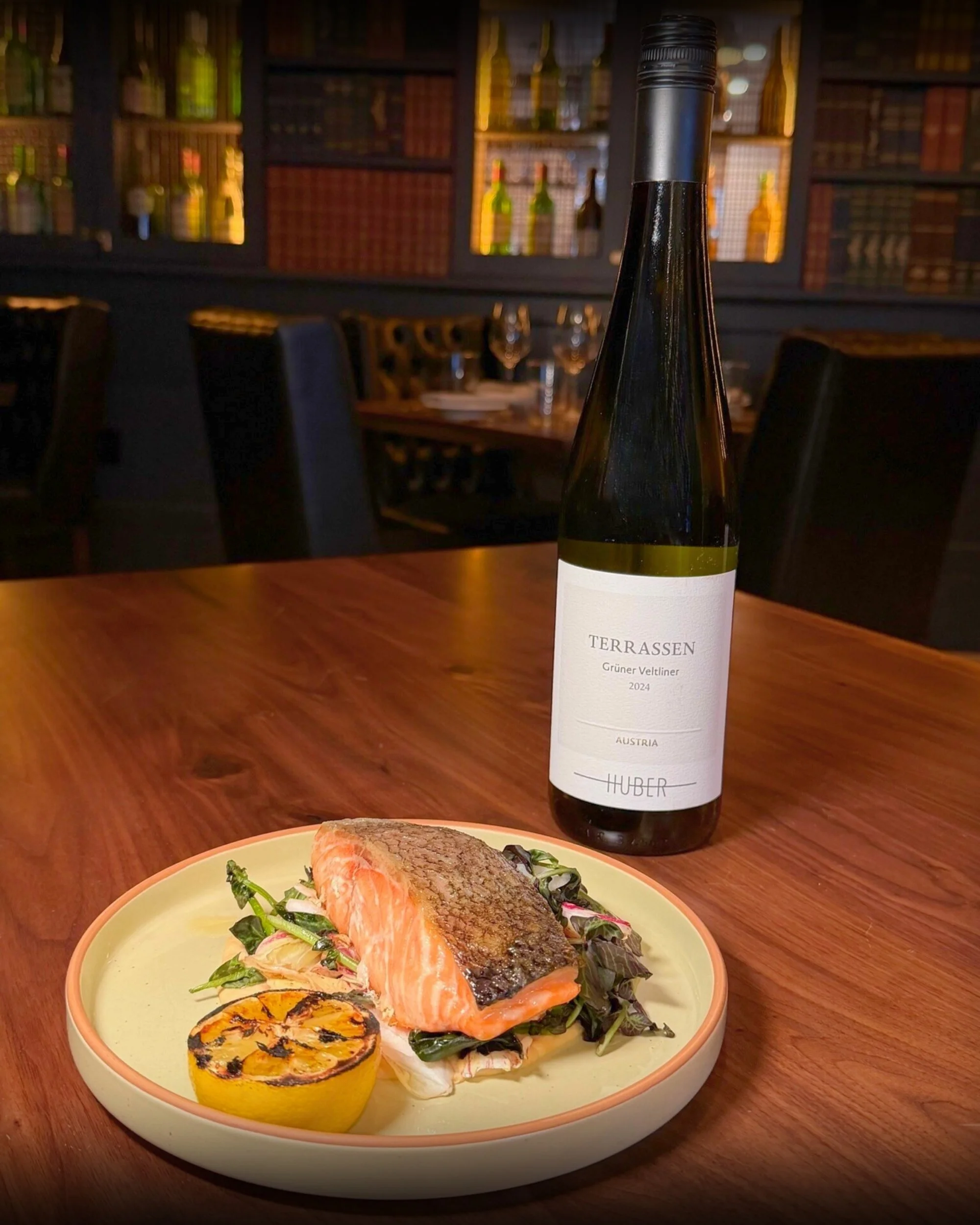 A plate with a grilled salmon fillet on a bed of greens and a grilled lemon half, placed on a wooden table. A bottle of 'Terrassen' Grüner Veltliner wine from Austria is next to the plate. The background shows a dimly lit restaurant with dark chairs and a bookshelf with bottles.