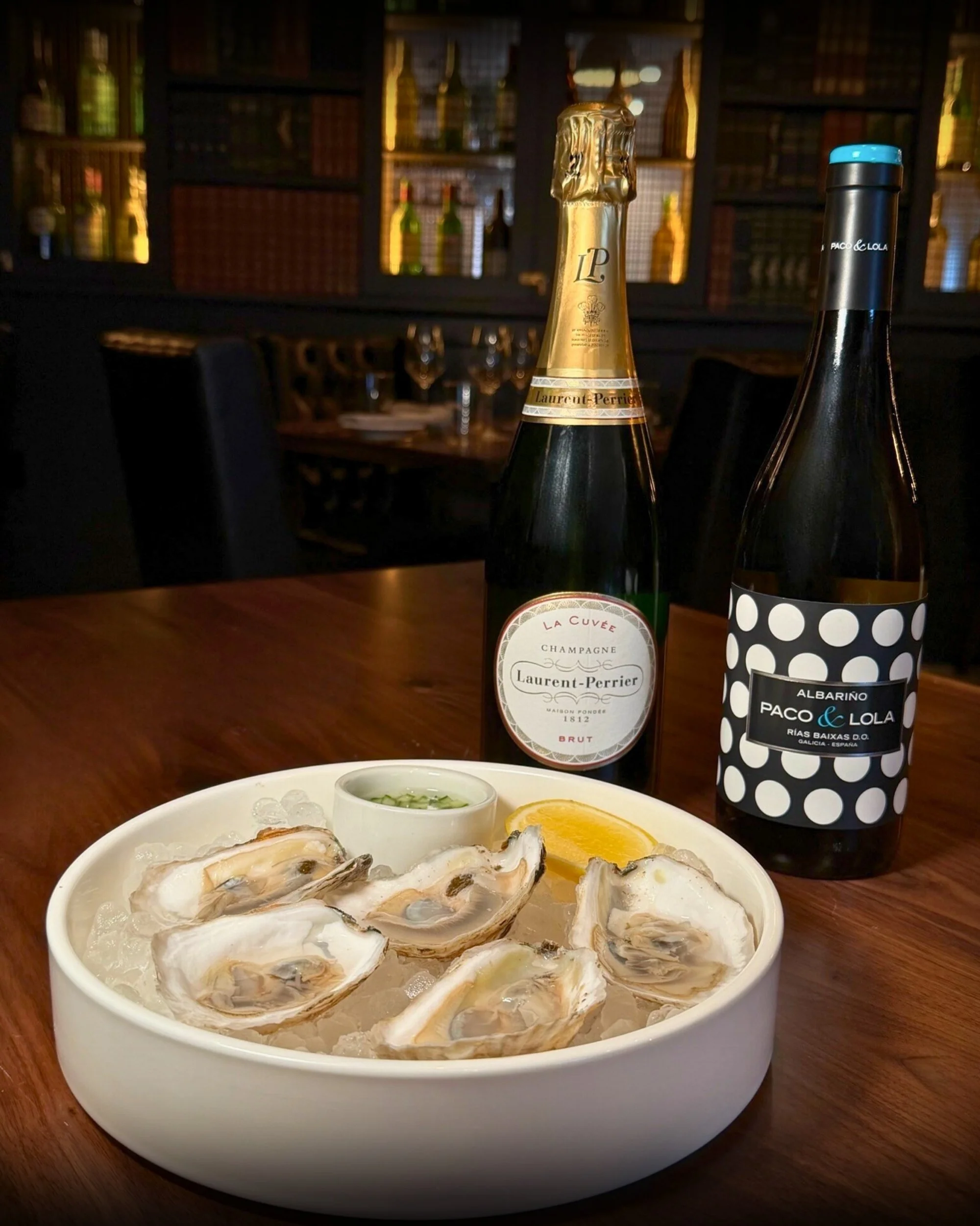 A white bowl of fresh oysters on ice with lemon wedge and dipping sauce, two bottles of sparkling wine and red wine on a wooden table in a dimly lit restaurant.
