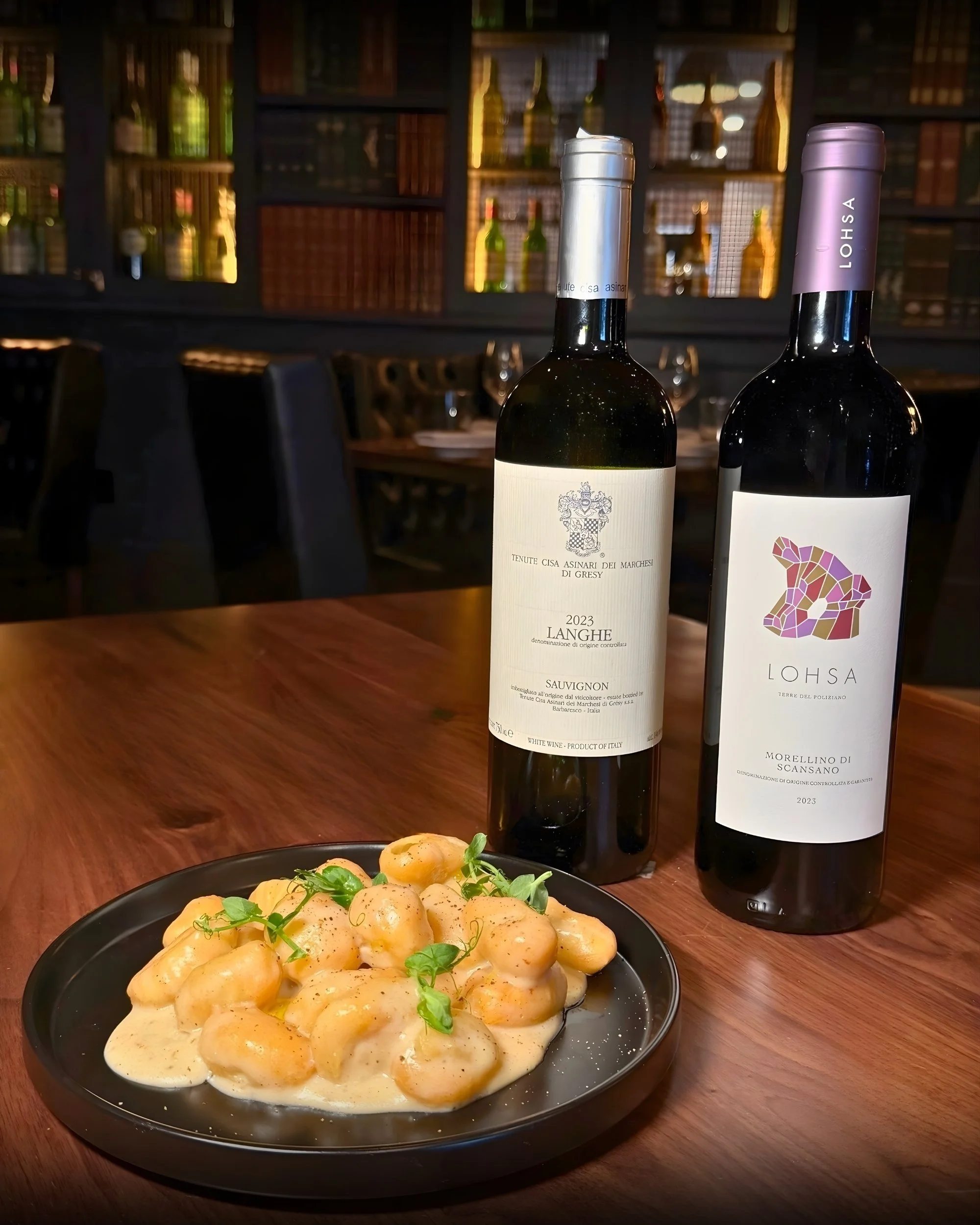 A black plate of gnocchi covered in a creamy white sauce with small green herbs on top, placed on a wooden table with two bottles of wine and a blurred background of a cozy restaurant.
