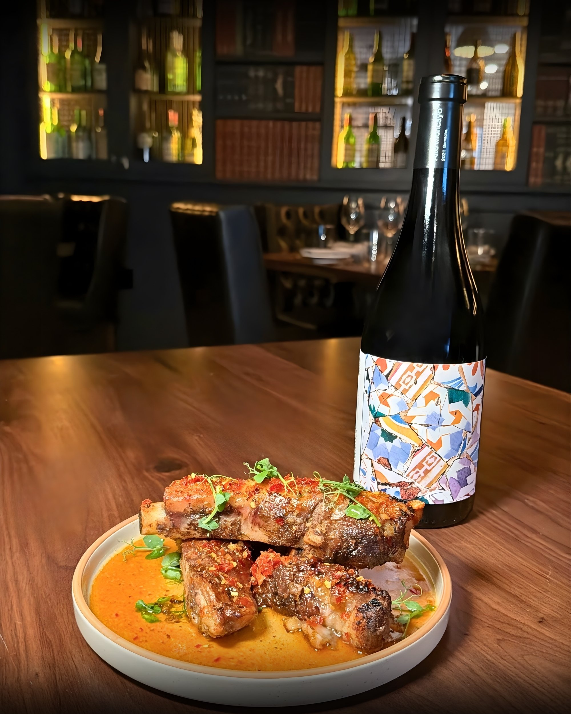 Dish of grilled meat ribs served with a sauce and garnished with herbs, with a bottle of wine in a restaurant setting.