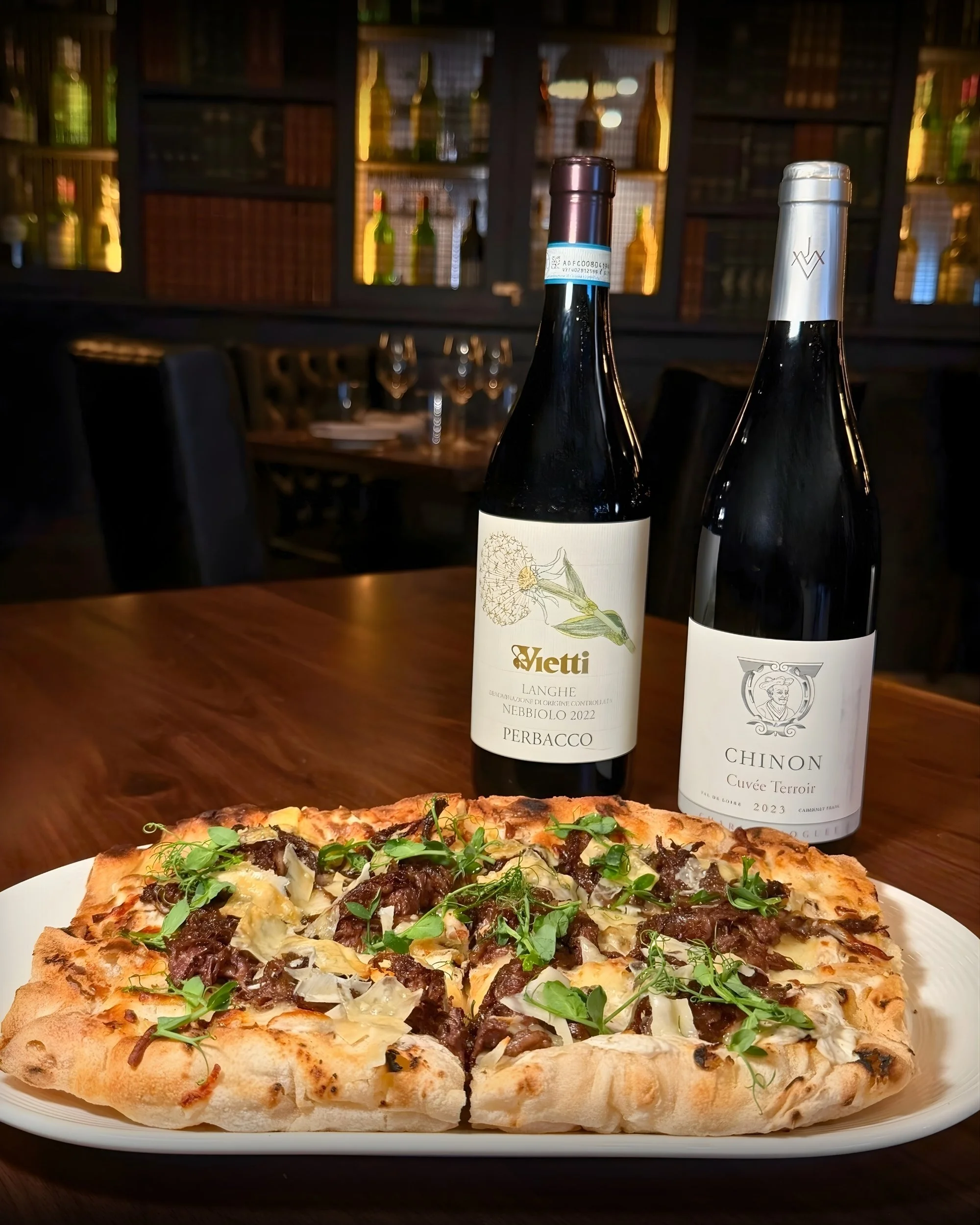 A rectangular pizza topped with mushrooms, meat, and fresh greens on a white plate, with two bottles of red wine in the background on a wooden table in a dimly lit restaurant.