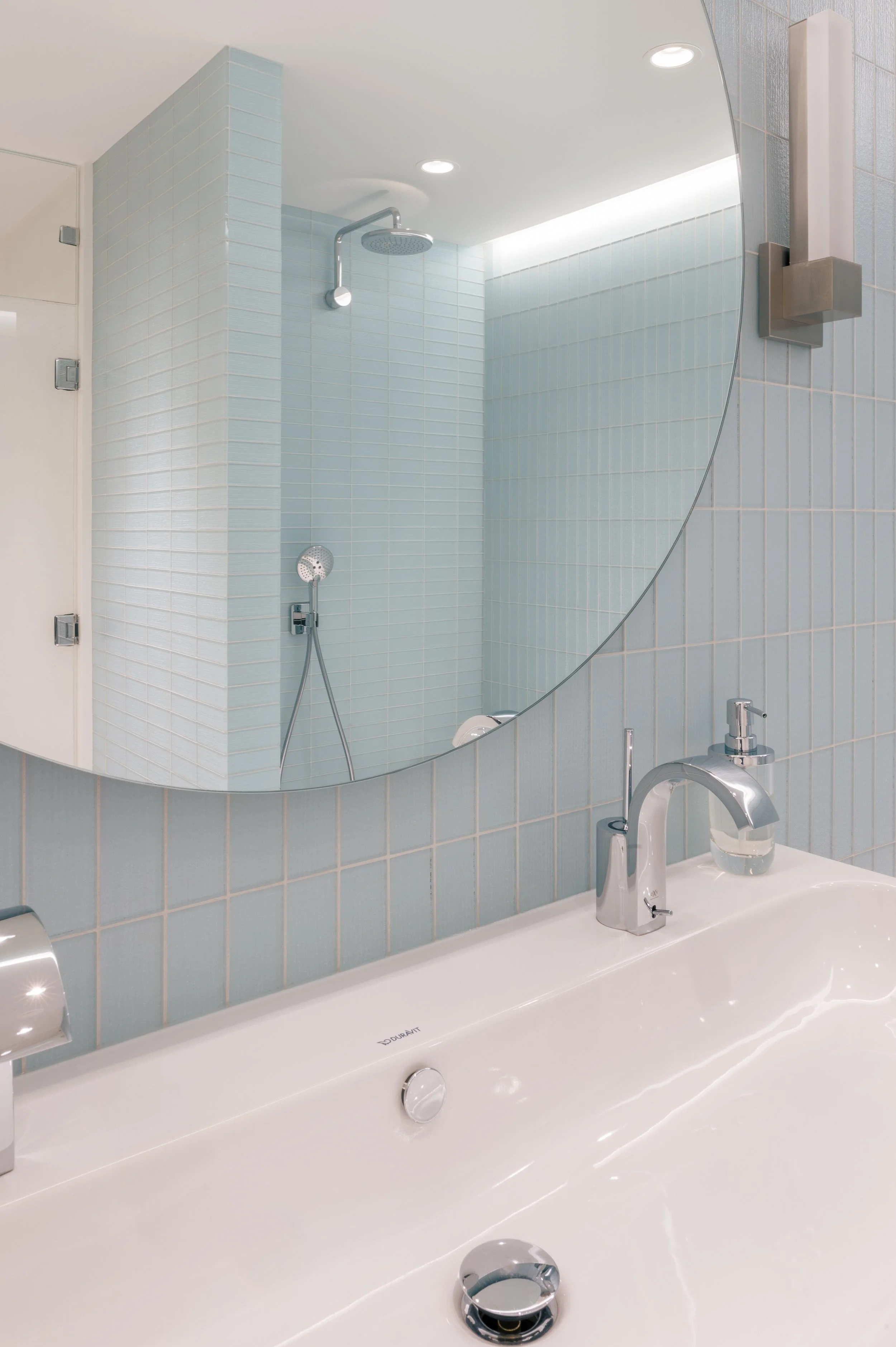 Modern bathroom with light blue tiled walls, a round mirror, silver faucets, and an overhead shower.