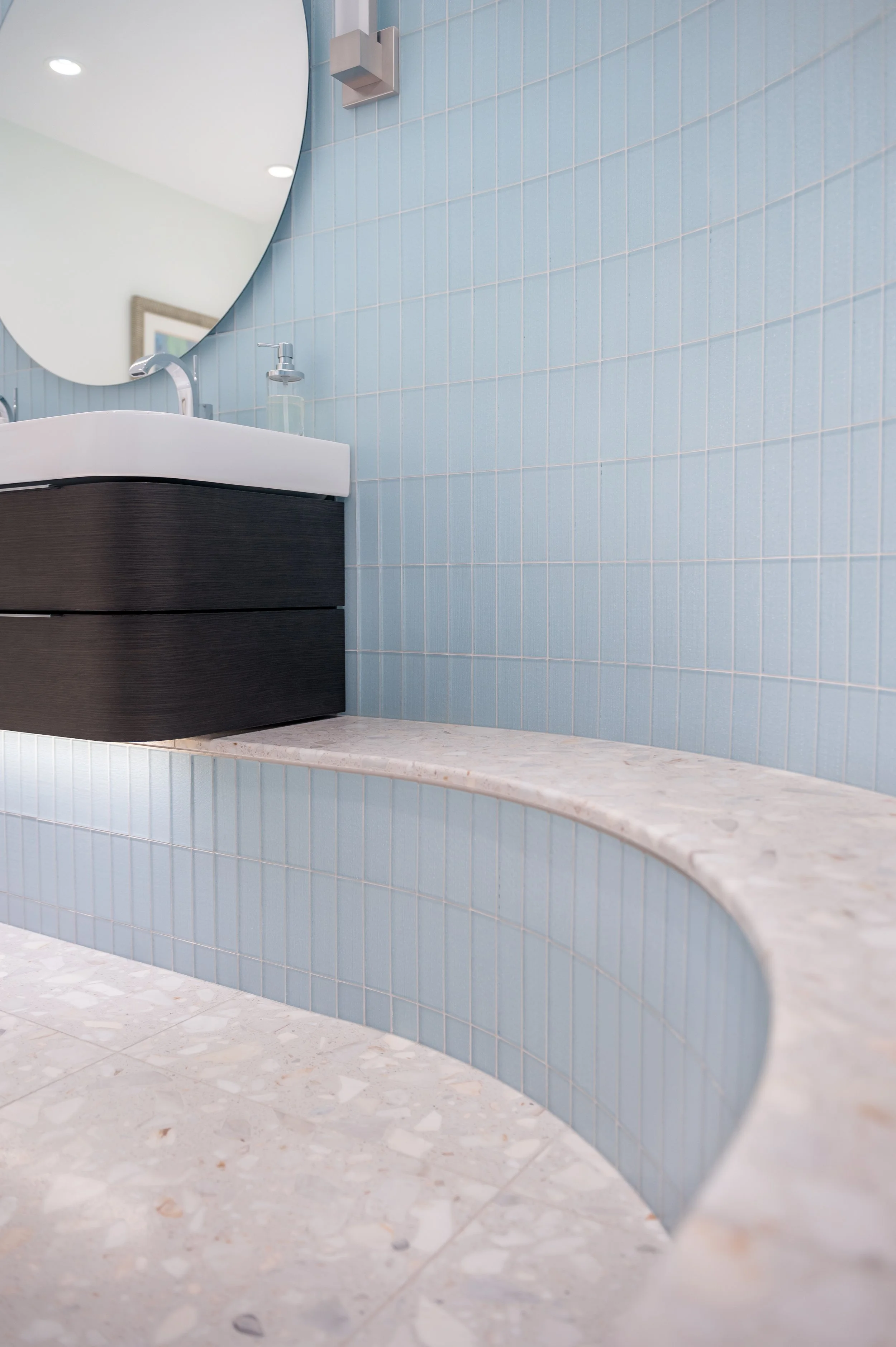 Modern bathroom with light blue tile walls, a circular mirror, a dark wood vanity, white sink, and marble-like tiled floor.